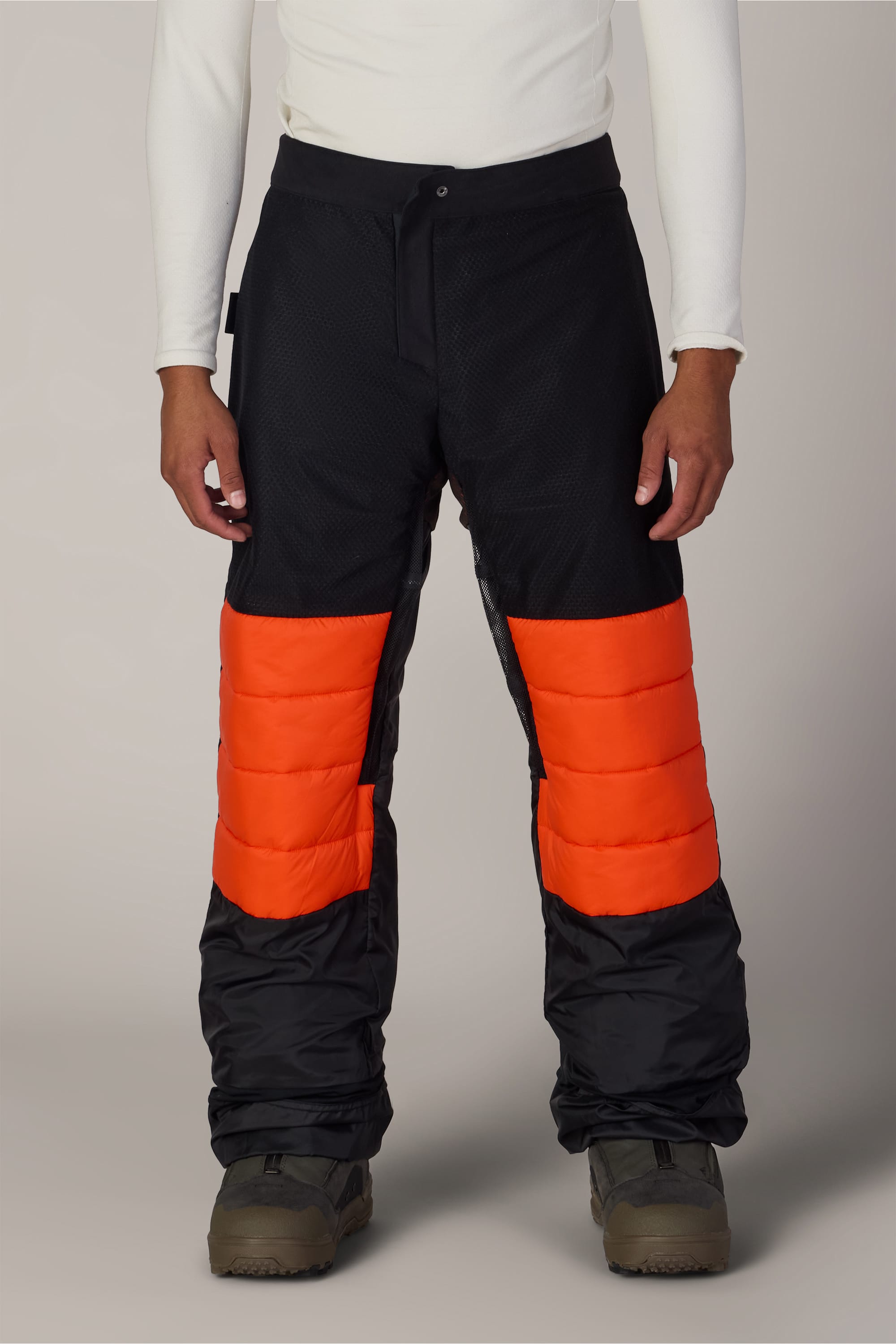 A person wearing the 686 Men’s Hydra™ Thermagraph® Pant by 686, featuring black nylon fabric with bright orange quilted knee panels, paired with a white long-sleeve shirt and brown boots, standing against a plain background.