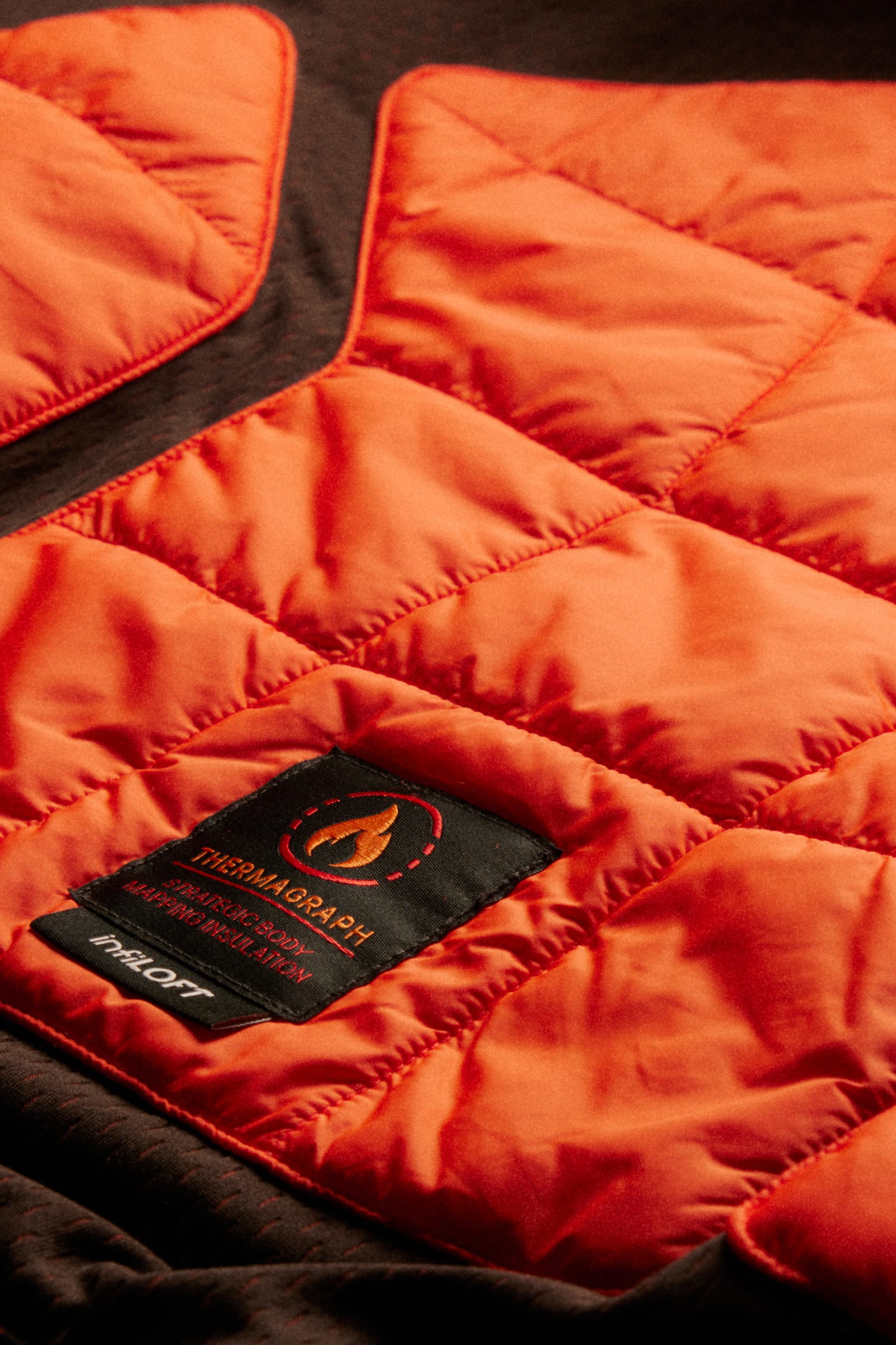 Close-up of an orange quilted jacket with a black label displaying the THERMAGRAPH® flame icon and additional text, with the 686 brand clearly visible at the bottom—a perfect complement to your favorite 686 Men’s Hydra™ Thermagraph® Pant or any men’s polyester pants.