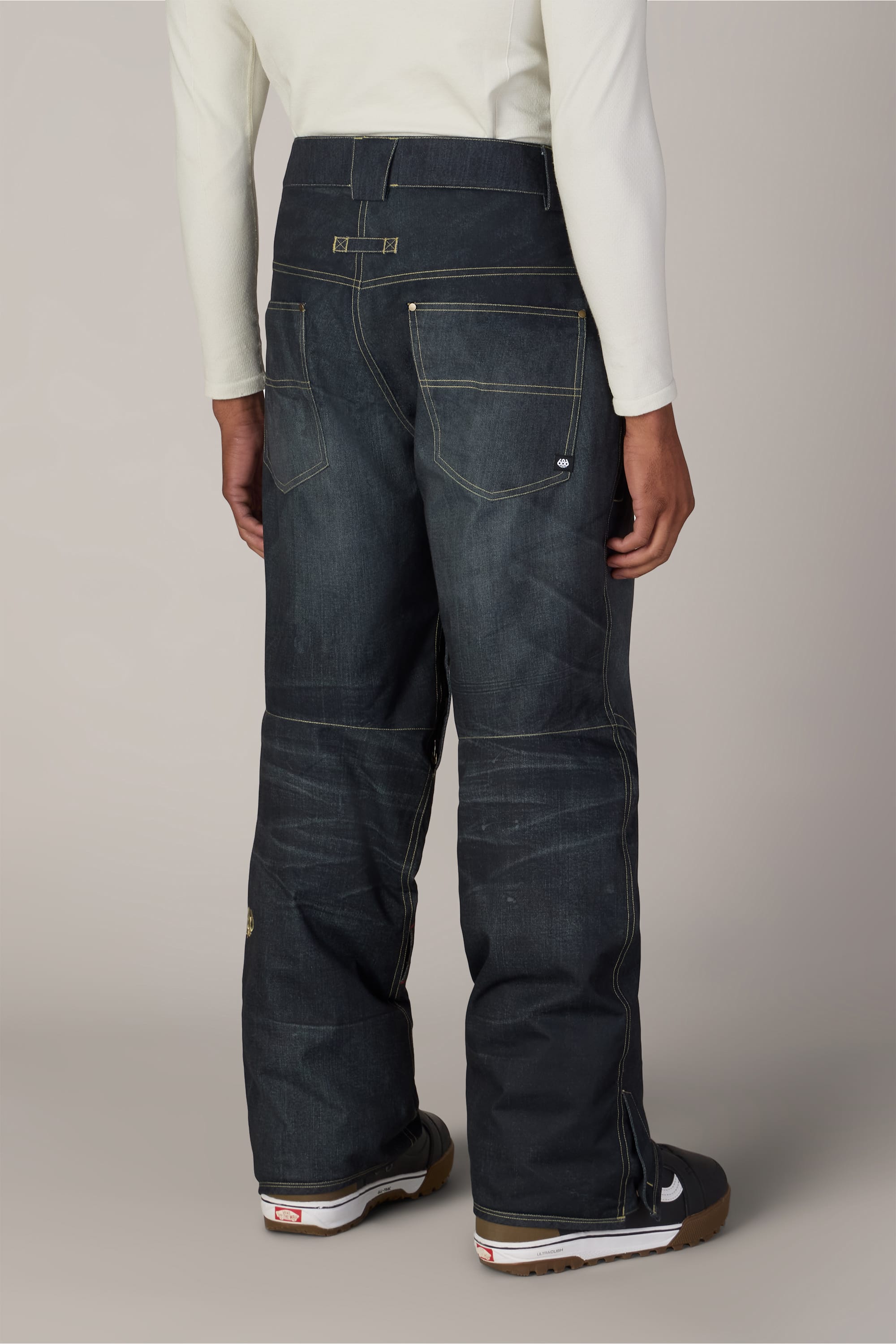 A person wearing 686 Men’s Deconstructed™ Denim Insulated Pant, featuring dark denim with yellow stitching, paired with a cream long-sleeve shirt, is shown from the back standing on a light-colored floor.
