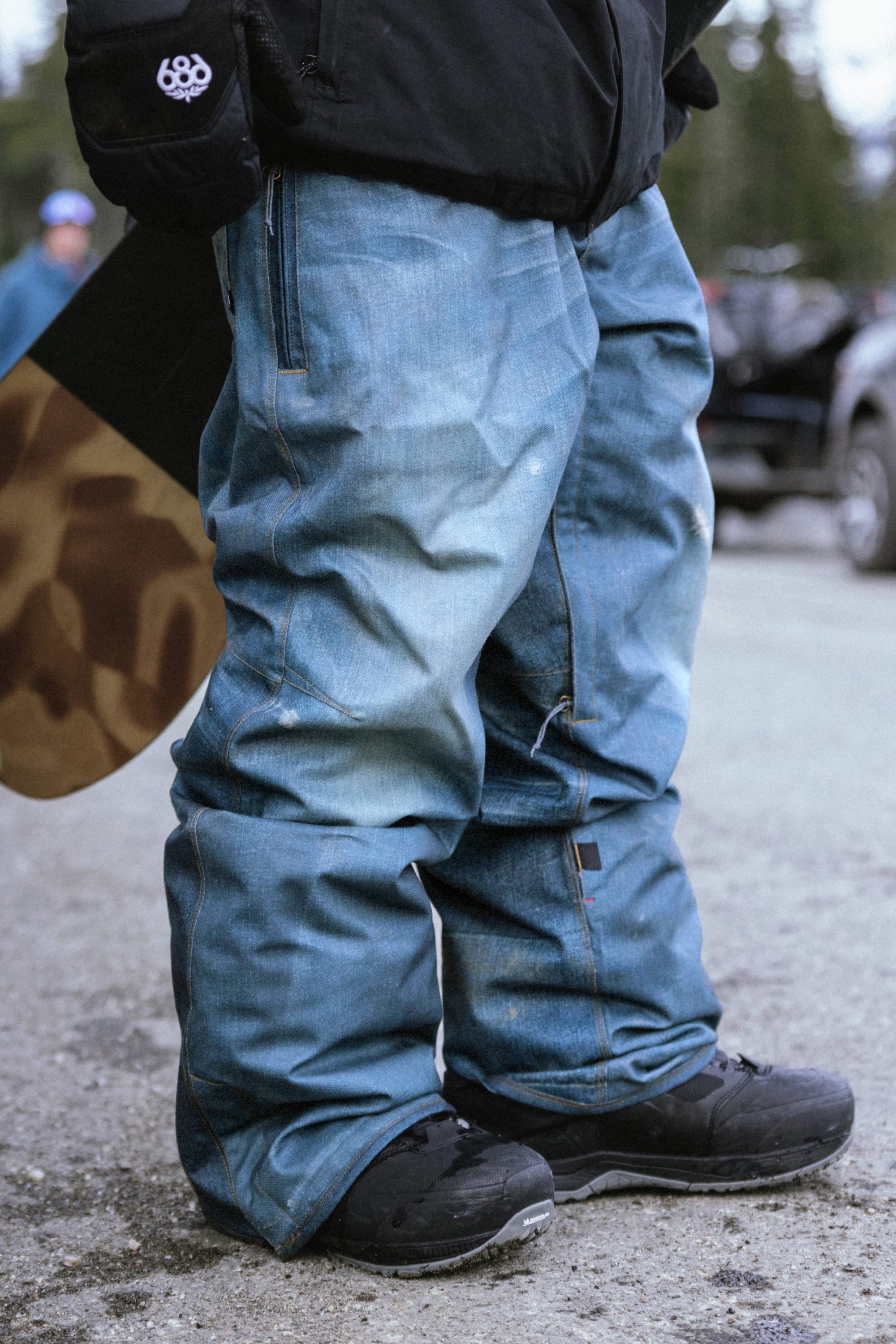 A person wearing the 686 Men’s Deconstructed™ Denim Insulated Pant and black snow boots stands on pavement, holding a snowboard. Trees and people are blurred in the background, suggesting a winter outdoor setting.
