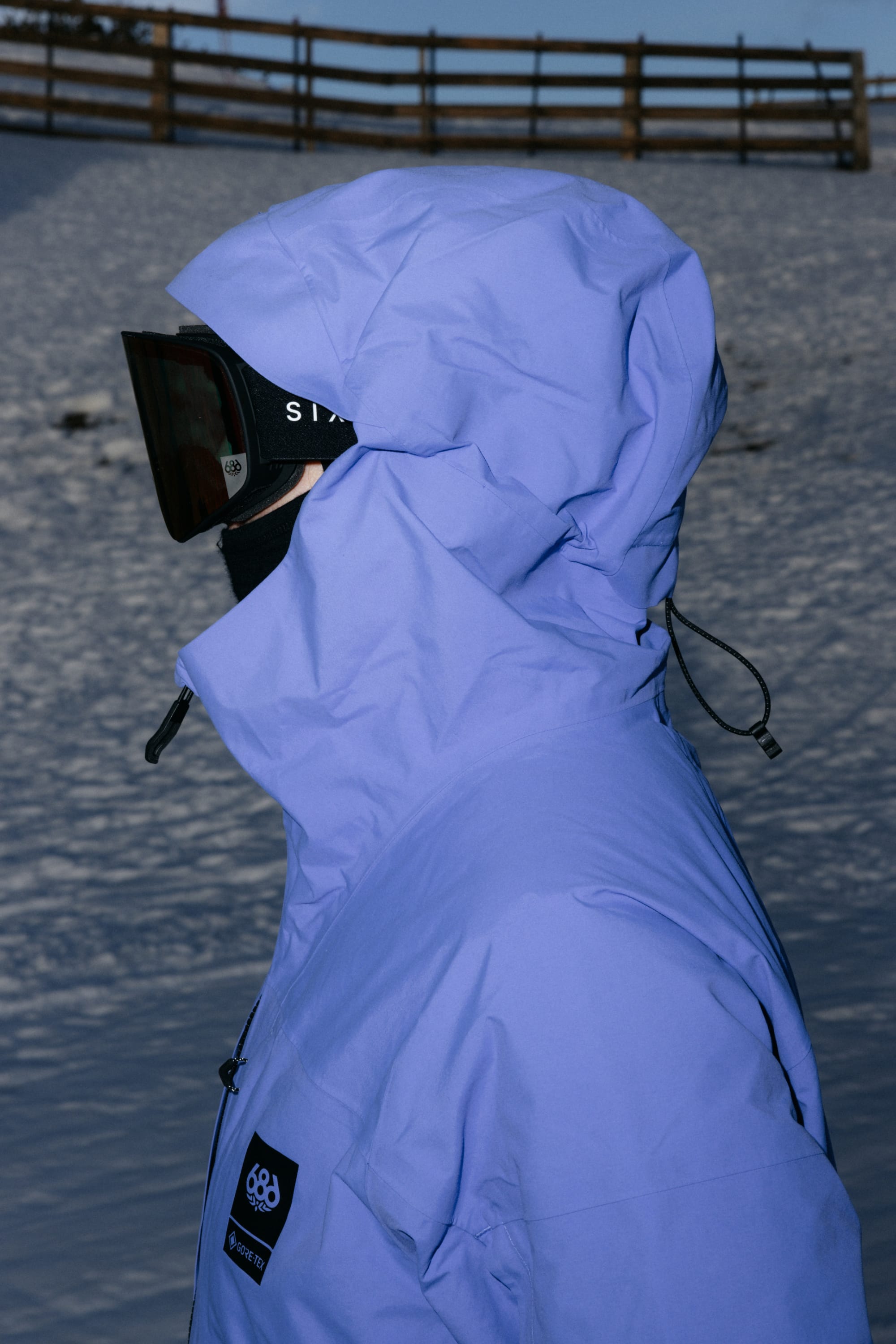 A person wearing the 686 Women’s GORE-TEX® Scheme Thermagraph® Jacket by 686, along with black ski goggles and a black face covering, stands in a snowy outdoor setting with a wooden fence in the background.