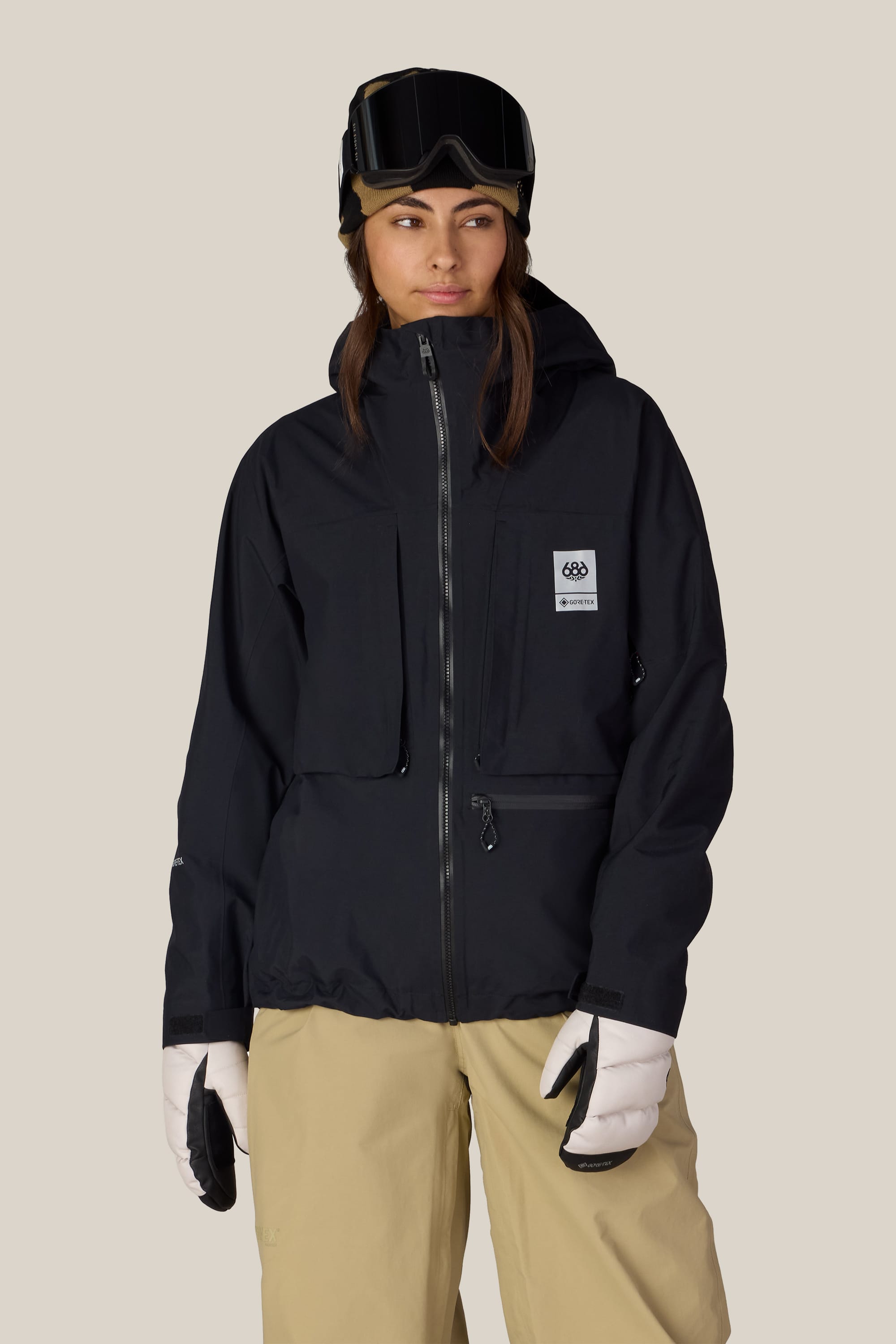 A person wearing the 686 Women’s GORE-TEX® Scheme Thermagraph® Jacket in black, paired with tan snow pants, white gloves, and ski goggles on their head stands against a plain light background.