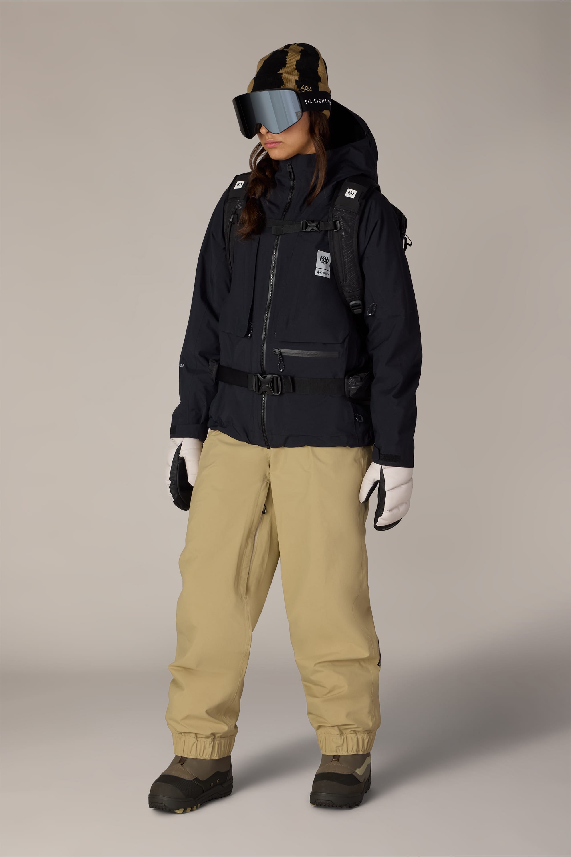 A person wearing the 686 Women’s GORE-TEX® Scheme Thermagraph® Jacket by 686, along with beige snow pants, gloves, boots, reflective ski goggles, and a camo hat stands against a neutral background, dressed for winter outdoor activities.