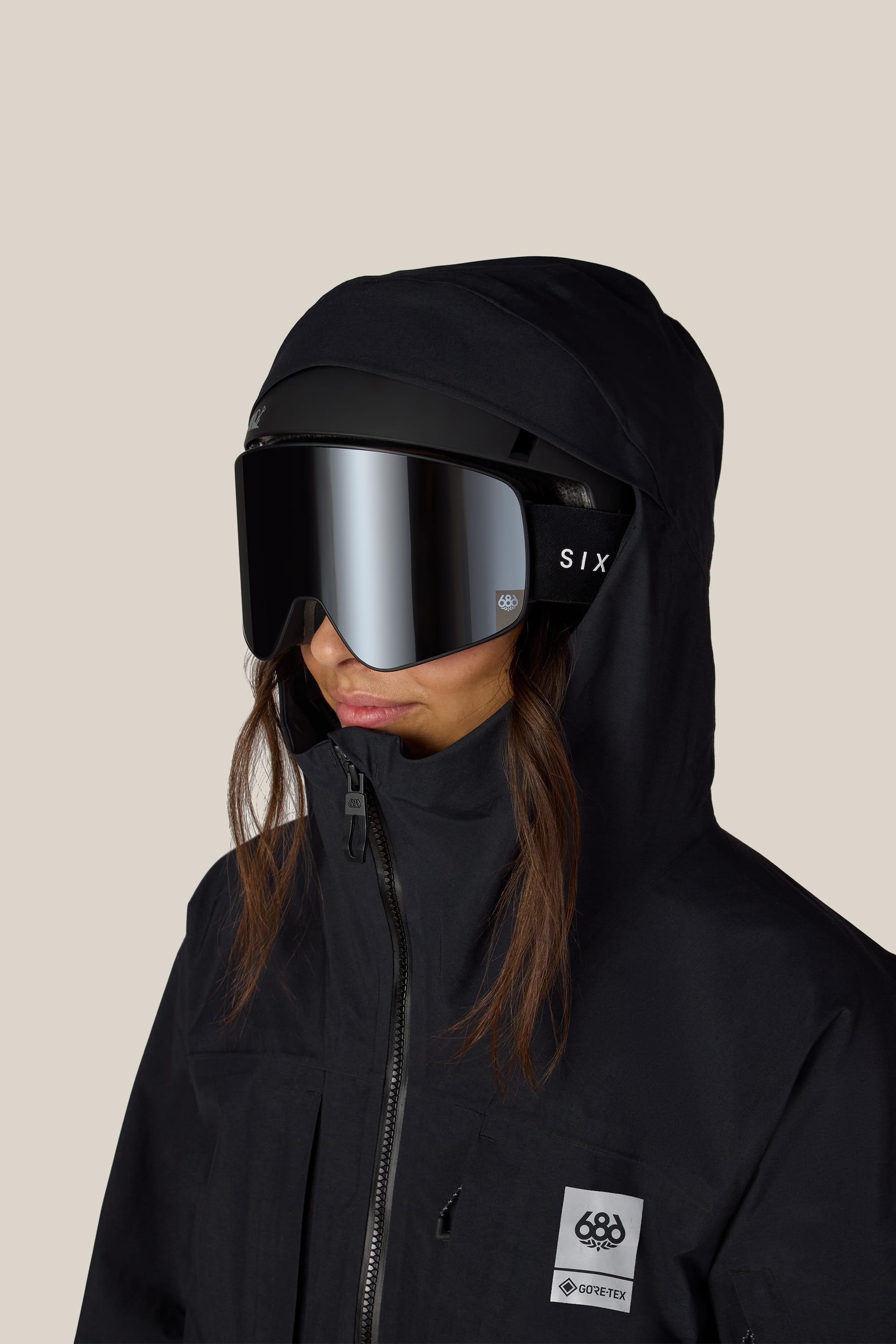A person is wearing the 686 Women’s GORE-TEX® Scheme Thermagraph® Jacket in black, along with a hood, reflective ski goggles, and black SIX-labeled headphones, facing left against a plain light background.
