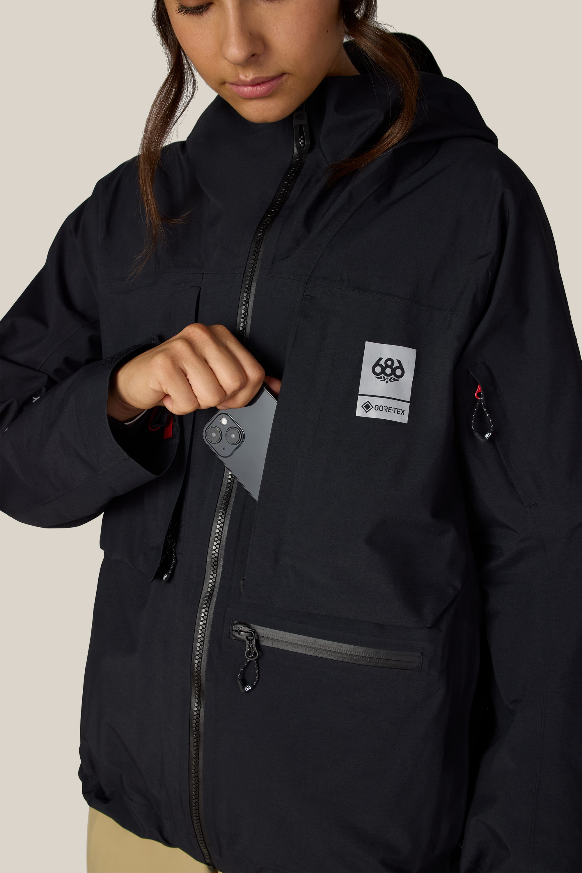 A person wearing a black 686 Women’s GORE-TEX® Scheme Thermagraph® Jacket places a black smartphone into the zippered left chest pocket, which features a visible 686 brand logo patch.