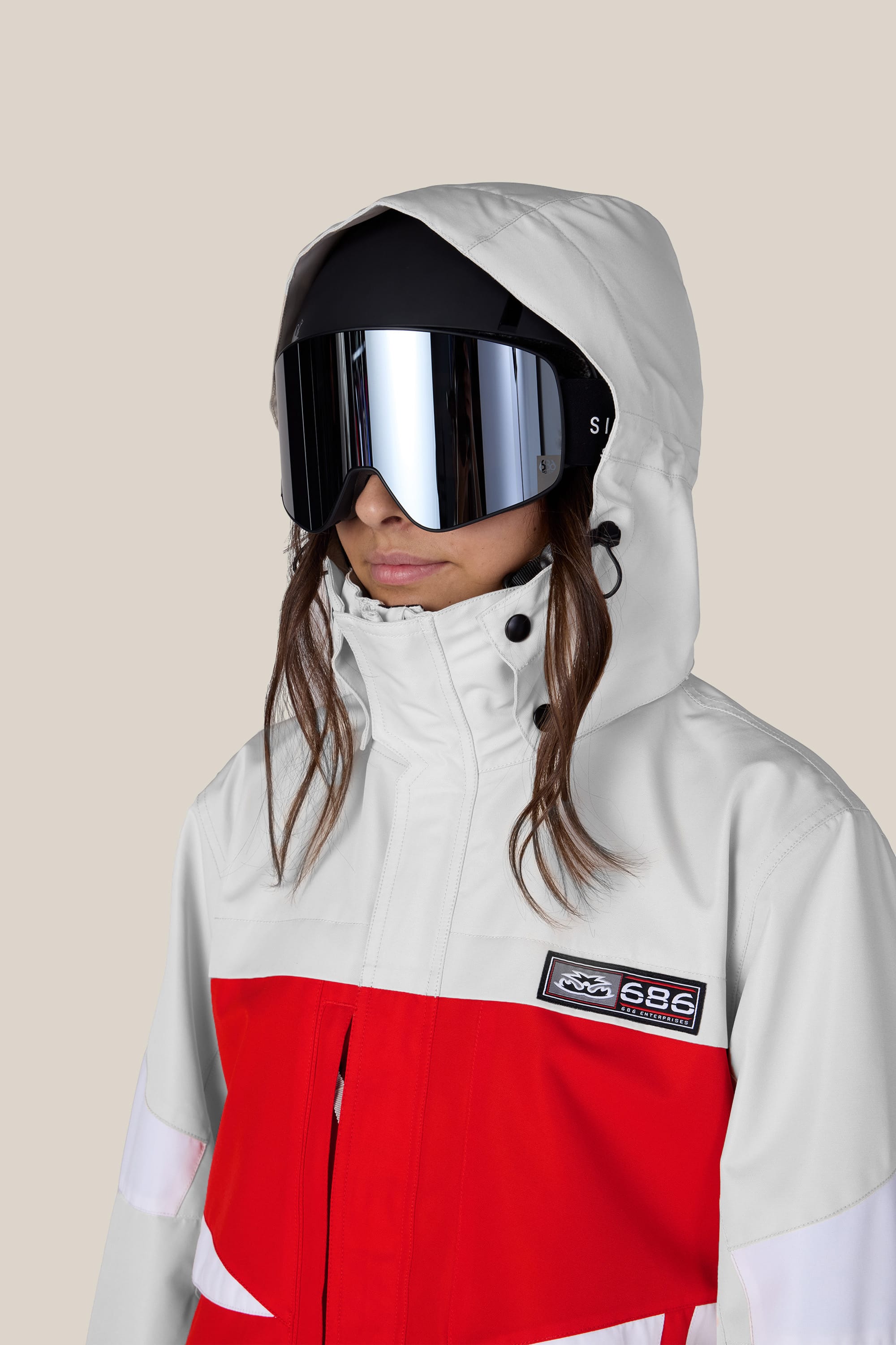 A person wearing the 686 Women’s Search Jacket in white, accented with red details, and large reflective ski goggles stands against a plain beige background.