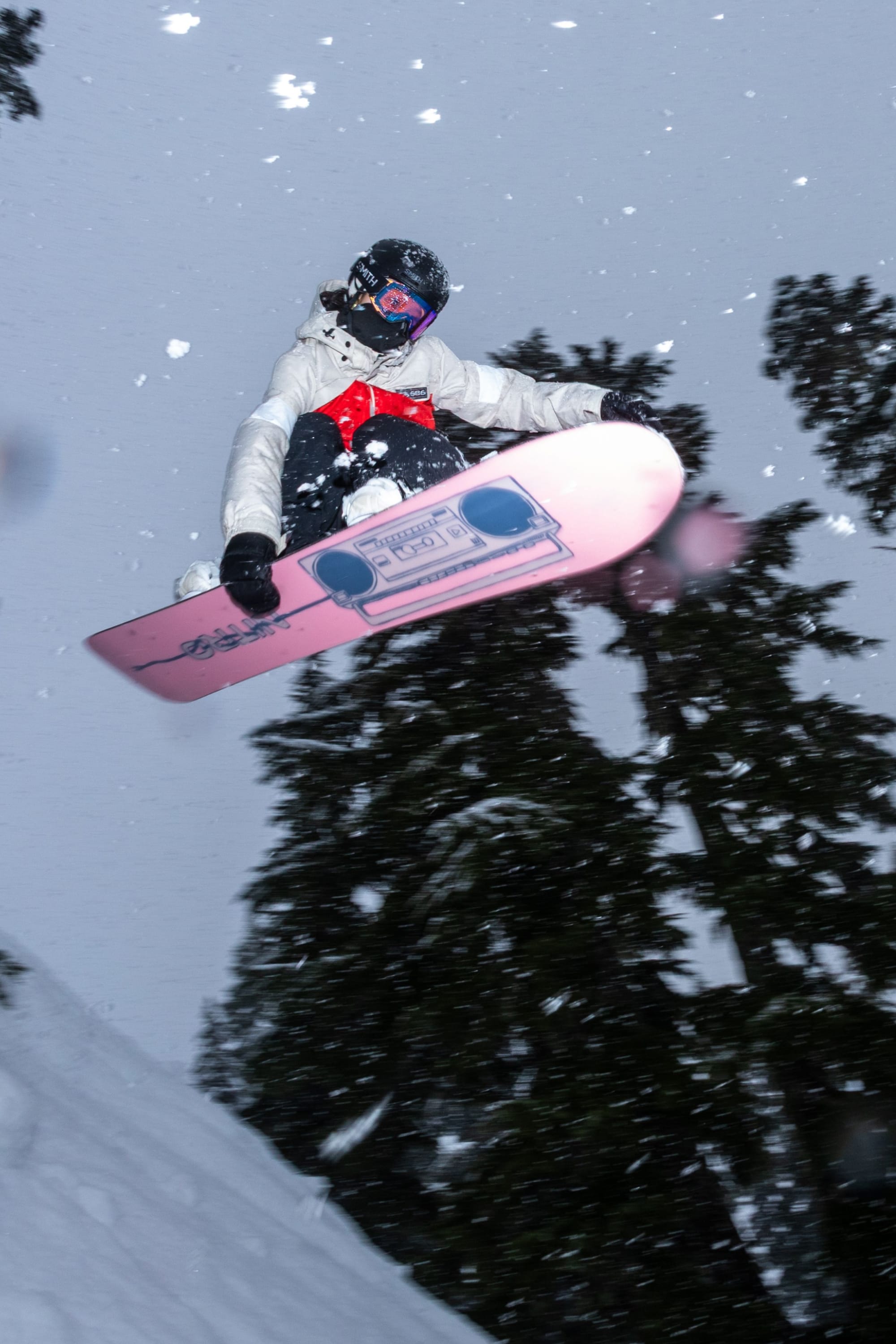 A snowboarder in winter gear catches air while jumping off a snowy slope surrounded by tall pine trees. Snowflakes swirl around her and her pink, graphic snowboard as she soars through the wintry landscape. She stays warm and dry in the 686 Women’s Search Jacket by 686, designed to keep her comfortable no matter how adventurous the ride gets.