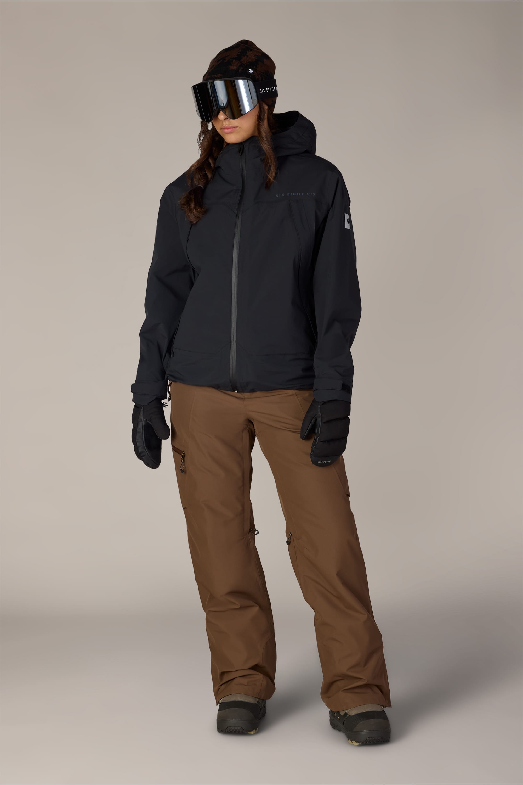 A person is wearing the 686 Women’s SMARTY® 3-in-1 Framework™ 3L Jacket by 686—a black polyester waterproof jacket—paired with brown snow pants, black ski gloves, black boots, and large ski goggles, standing against a plain light background.