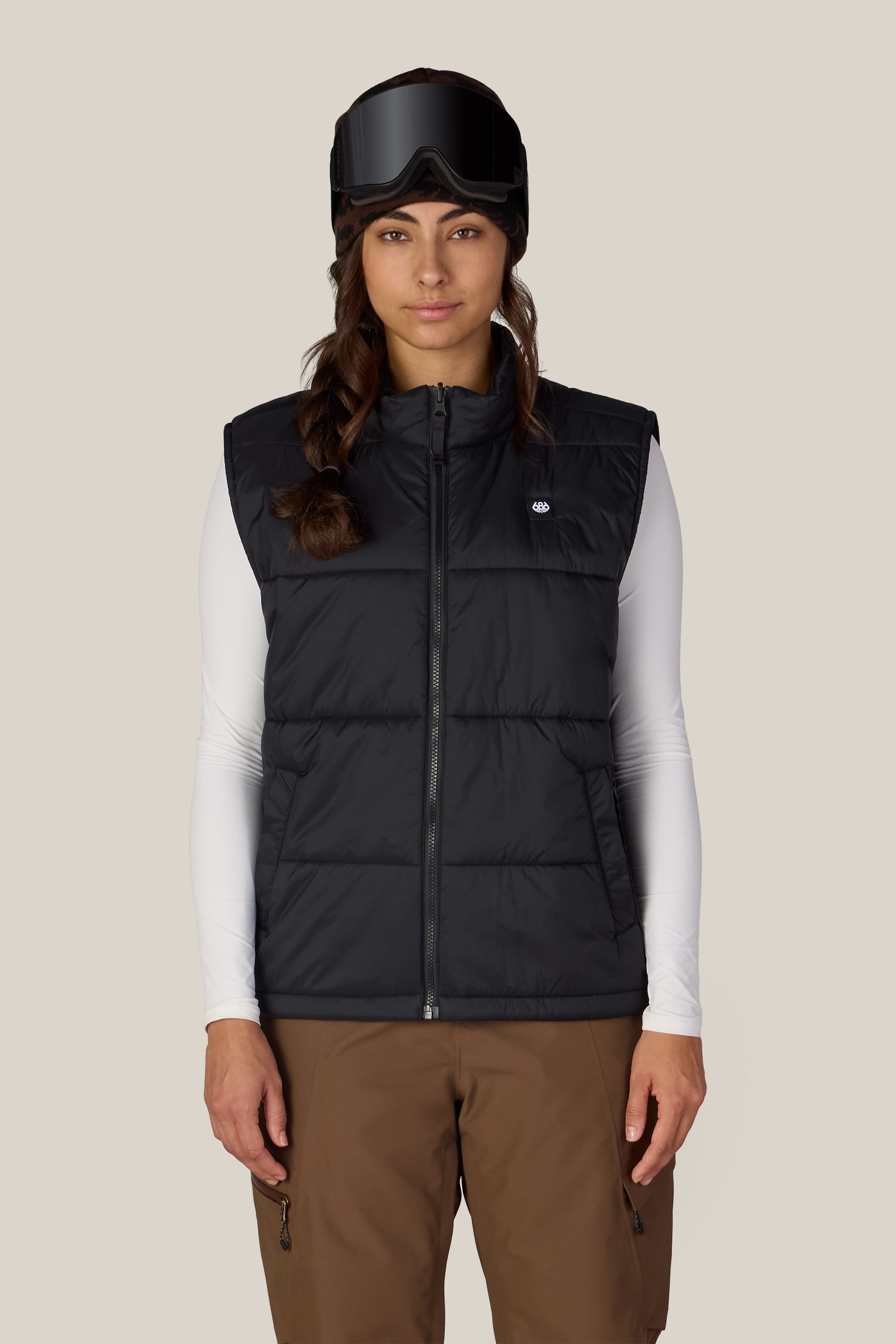 A woman stands facing forward against a plain background, styled in the 686 Women’s SMARTY® 3-in-1 Framework™ 3L Jacket layered under a black puffer vest, paired with ski goggles, a white long-sleeve shirt, and brown pants—effortlessly highlighting this versatile and waterproof jacket by 686.