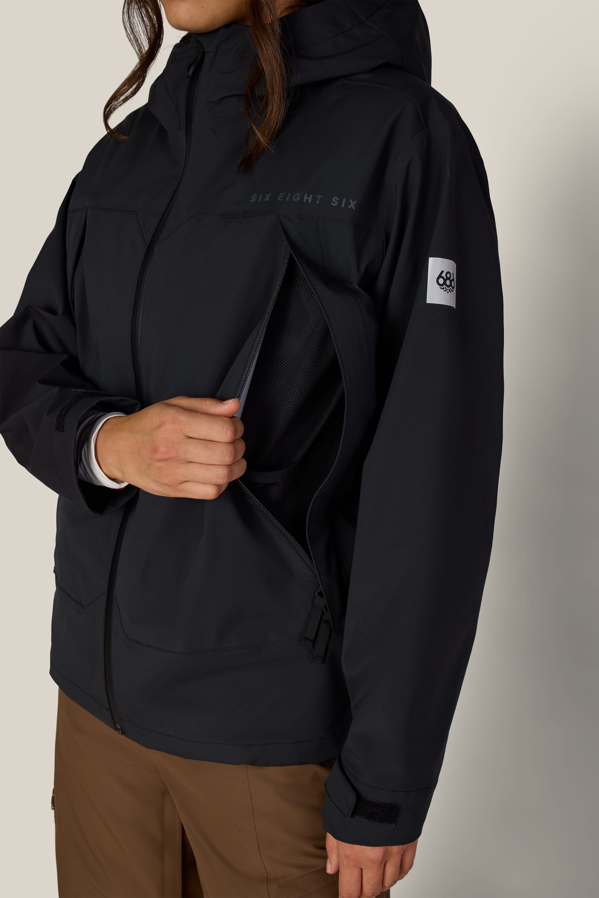 A person is wearing the 686 Women’s SMARTY® 3-in-1 Framework™ 3L Jacket in black with brown pants, unzipping a ventilation pocket on the side; this polyester waterproof jacket by 686 features a small white patch on the sleeve and a subtle logo on the chest.