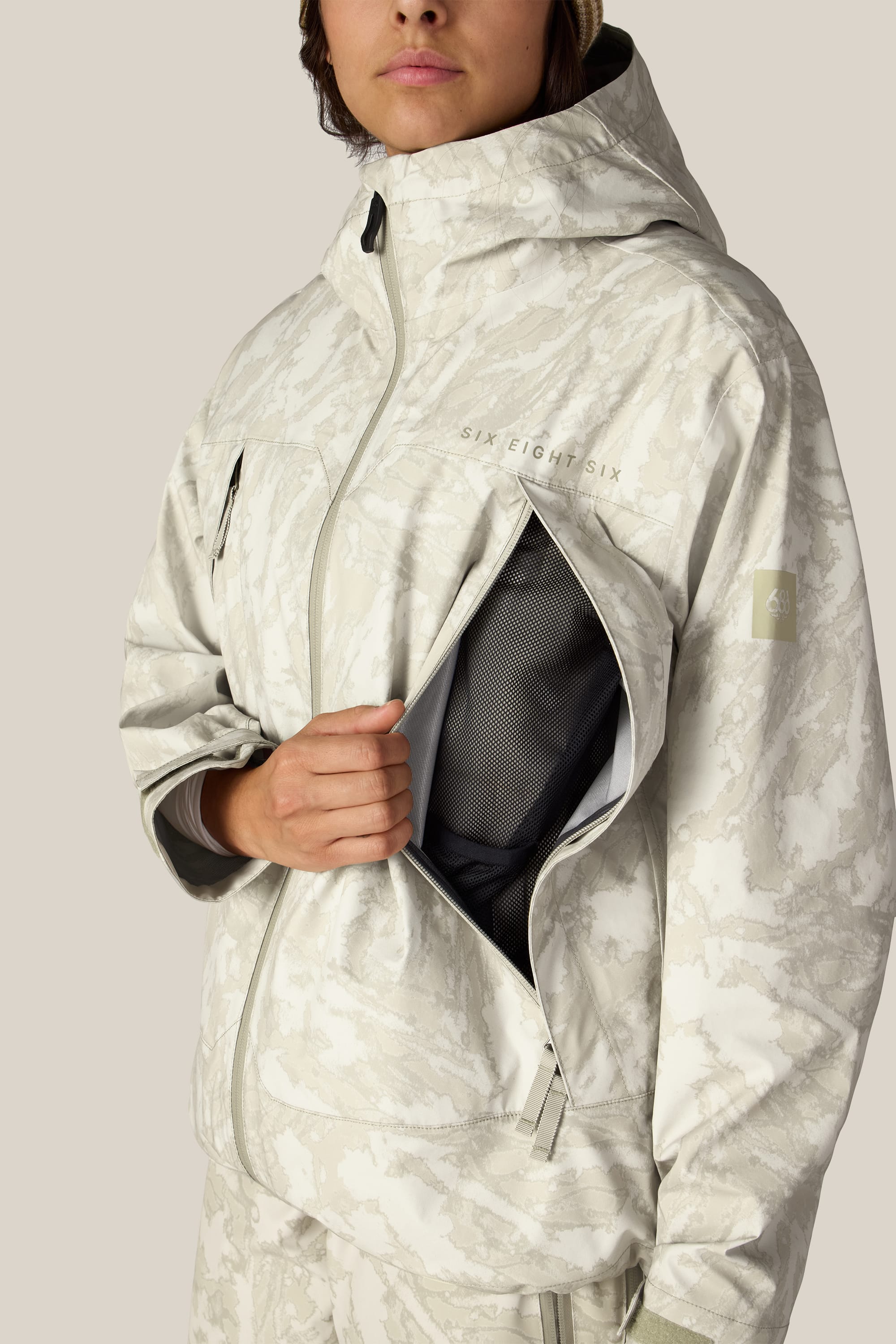 A person wearing the 686 Women’s SMARTY® 3-in-1 Framework™ 3L Jacket, a light-colored, patterned woven jacket with a hood, unzips a large chest pocket to reveal the mesh lining inside. Only the lower part of their face is visible against a plain beige background.