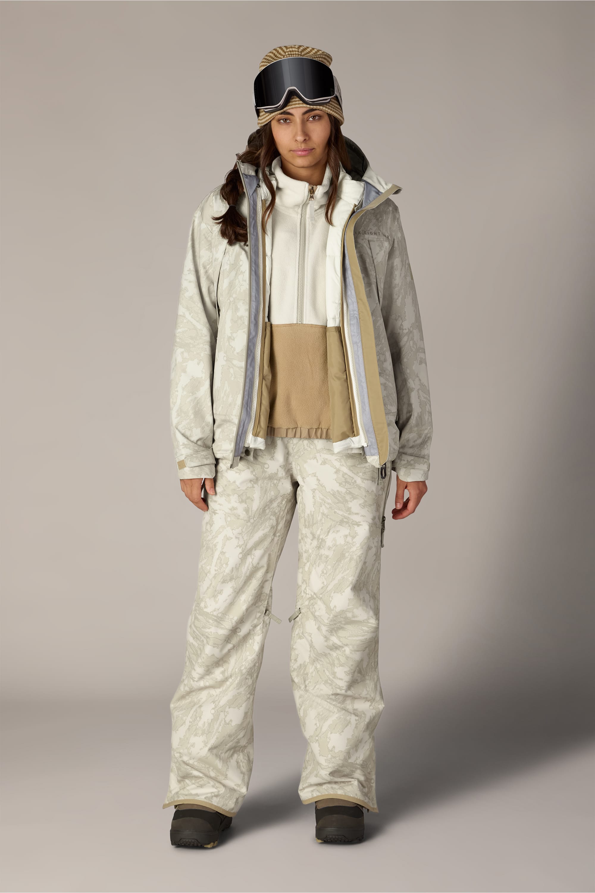 A person stands facing forward against a plain background wearing the 686 Women’s SMARTY® 3-in-1 Framework™ 3L Jacket in beige and white, paired with matching pants, a hoodie, ski goggles on their head, and black boots.