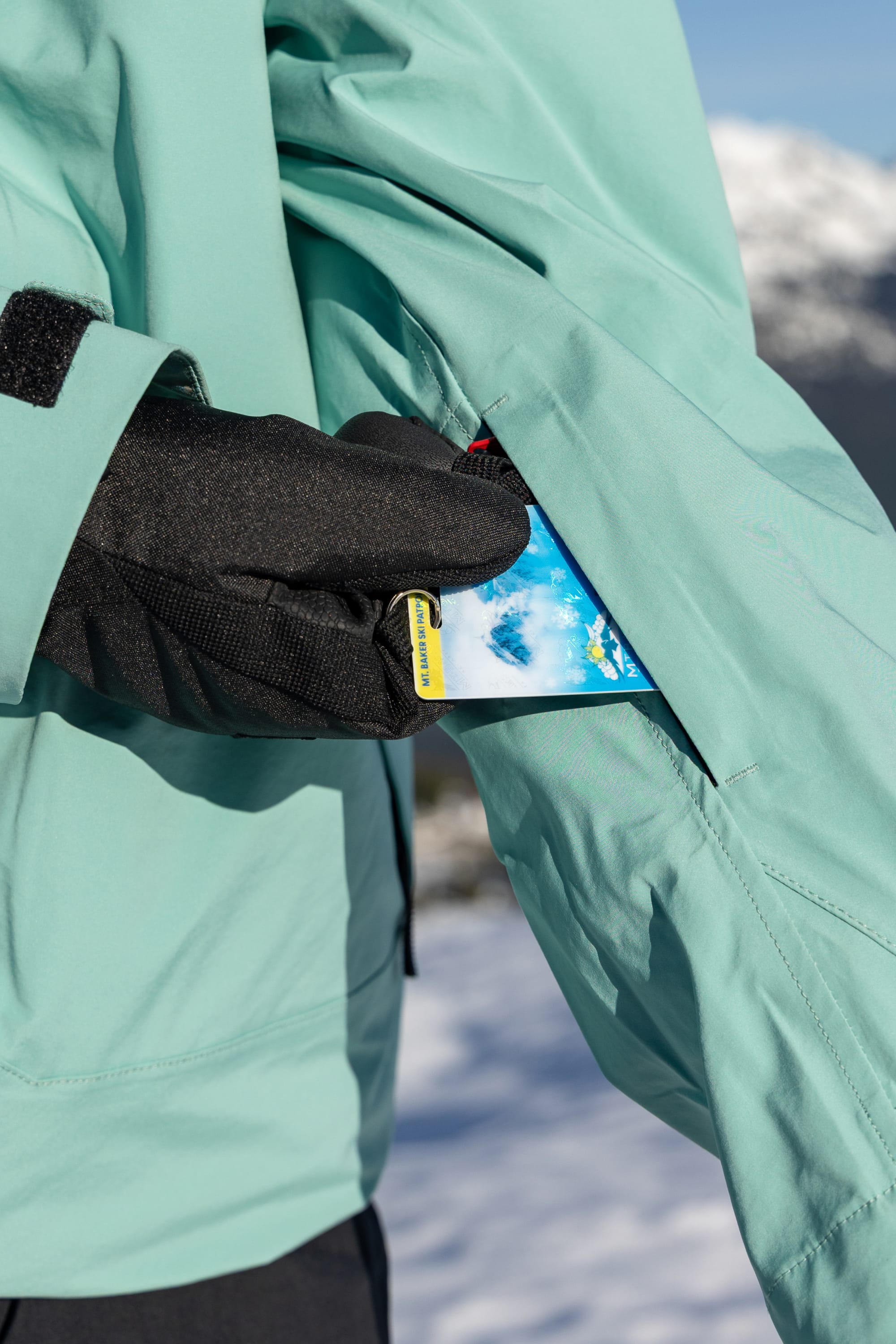Wearing black gloves and the 686 Women’s SMARTY® 3-in-1 Framework™ 3L Jacket, a person slips a ski pass into the jacket’s zippered sleeve pocket, with snowy mountains softly blurred in the background.