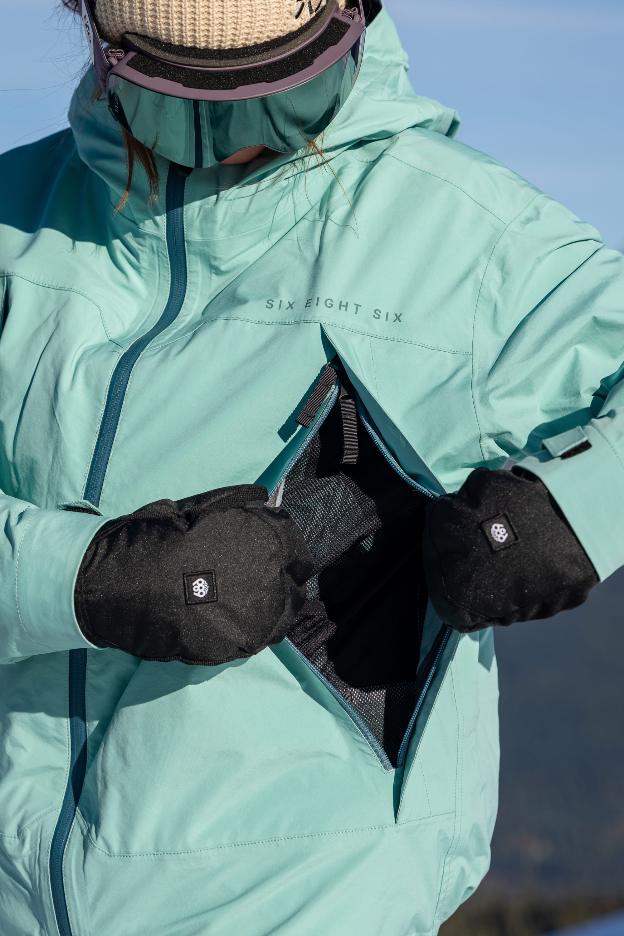 Wearing the 686 Women’s SMARTY® 3-in-1 Framework™ 3L Jacket in teal and black gloves, a person opens a zippered chest pocket outdoors. Their face is partially obscured by goggles and a tan beanie.