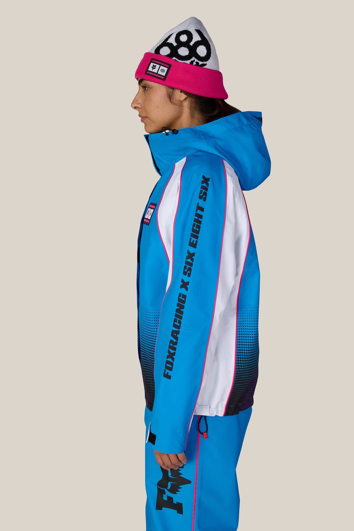 A person wearing a pink and white 686 beanie and a 686 Women’s Fox Racing 97 Shell Jacket and matching pants.