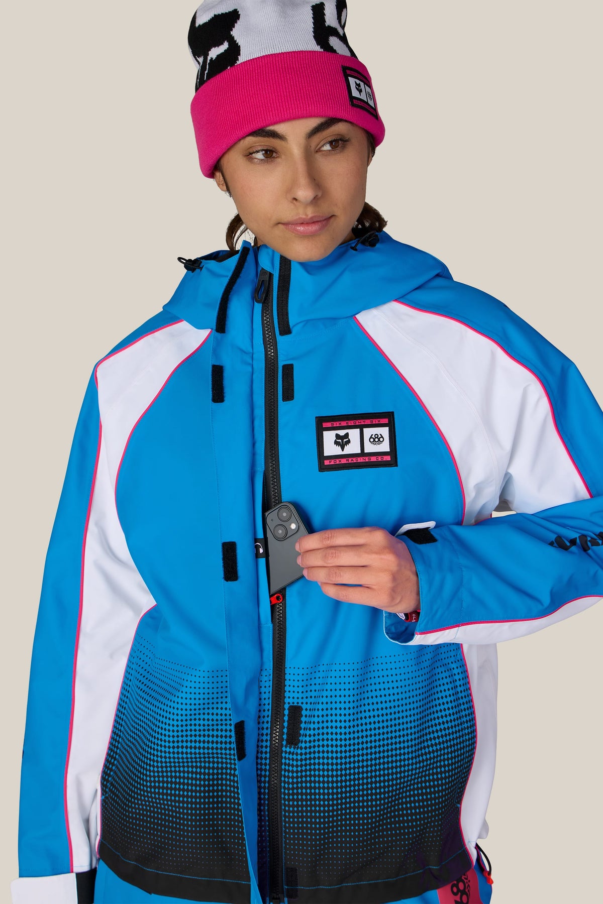 A person wearing a blue, white, and pink 686 Women’s Fox Racing 97 Shell Jacket and a pink beanie, pulling a smartphone from a chest pocket.