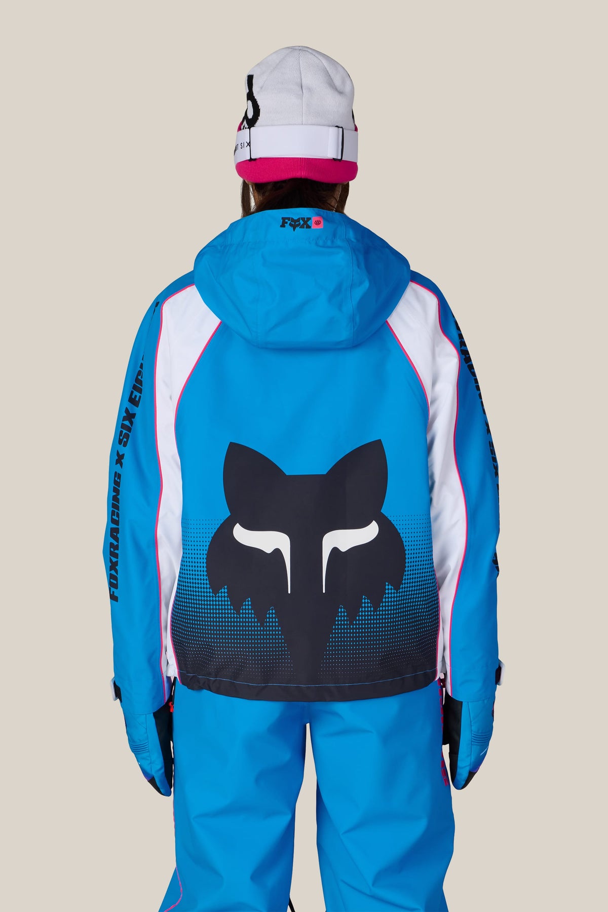 A person seen from behind wears a 686 Women’s Fox Racing 97 Shell Jacket with a large fox logo on the back, matching blue snow pants, and a white and pink beanie.