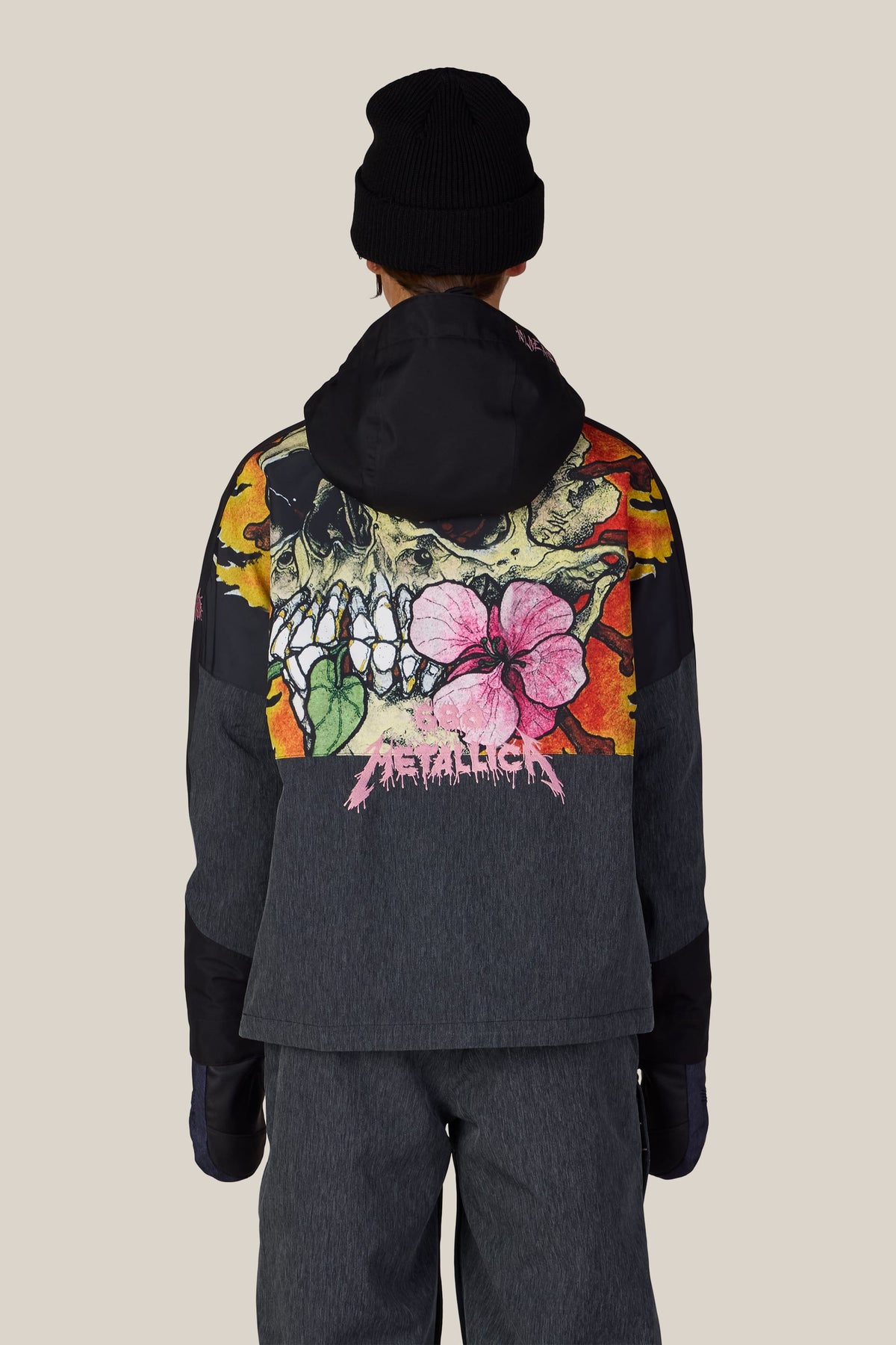 Back view of a person wearing a black beanie and the 686 Women’s Metallica Burning Skull Insulated Jacket, a dark gray waterproof jacket featuring a large graphic of a skull, flames, and a pink flower with the Metallica logo in pink.