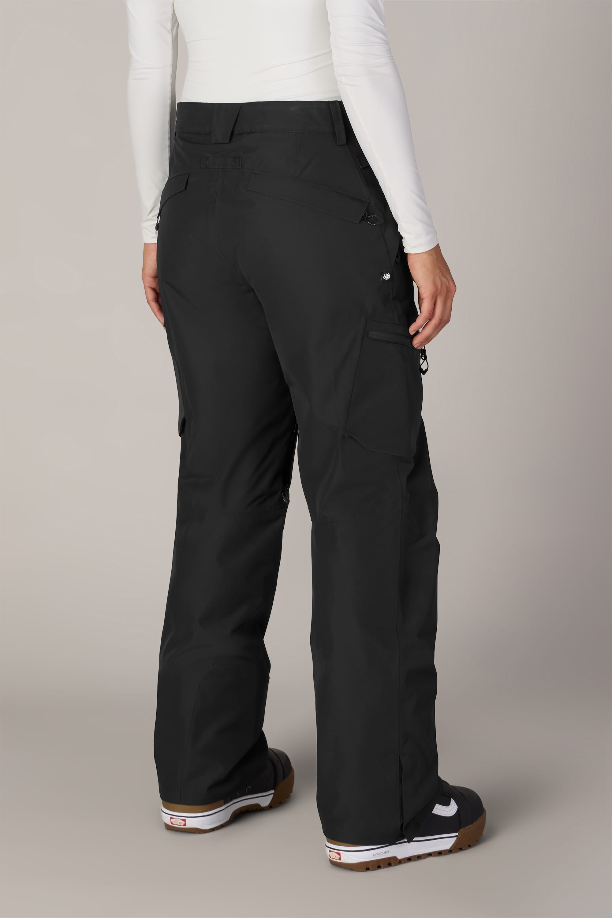 A person wearing the 686 Women’s Geode Thermagraph® Pant by 686 in black, paired with a white long-sleeve top and black sneakers with brown soles, stands facing away from the camera on a plain background.