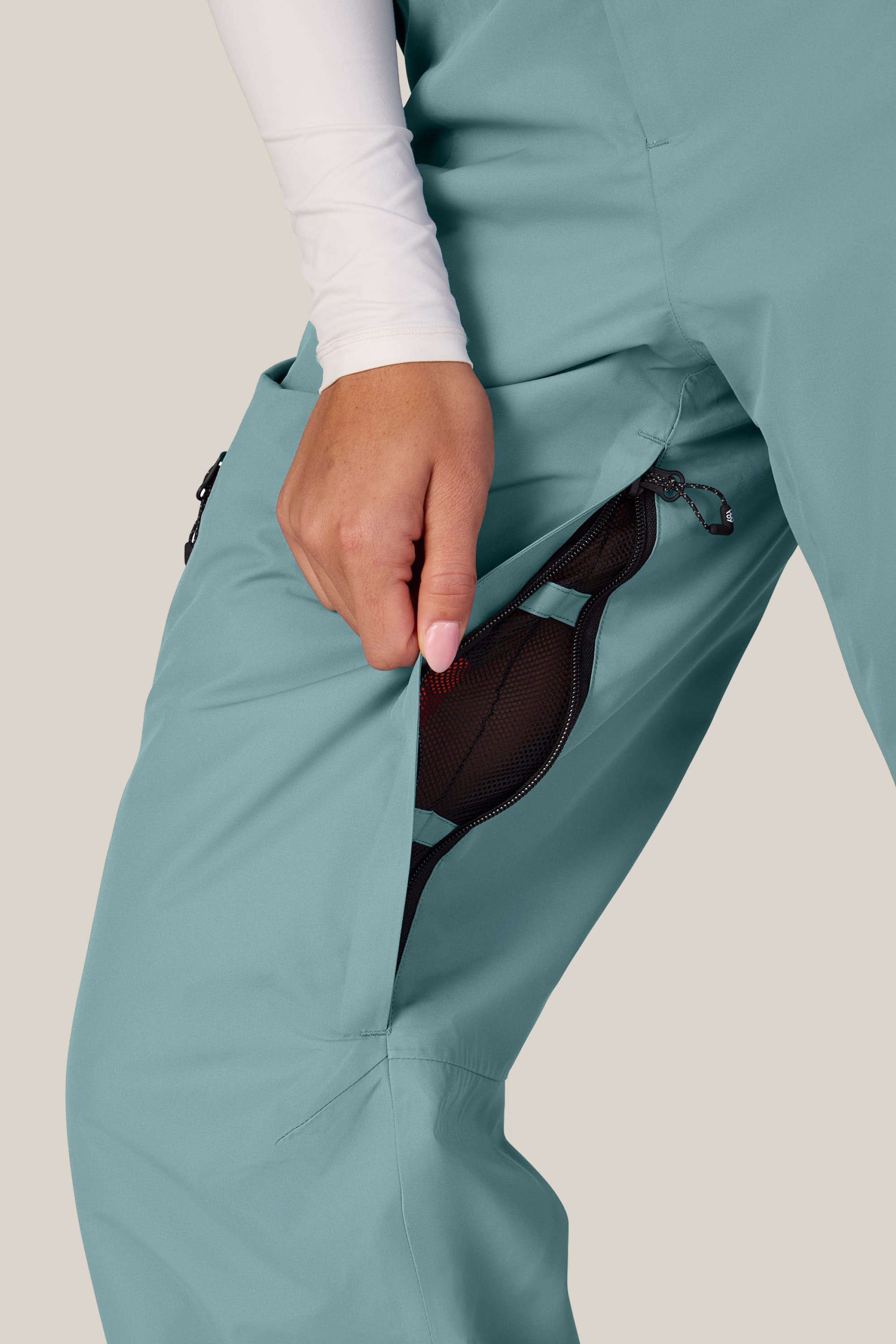 A person wearing the 686 Women’s Geode Thermagraph® Pant in teal has the zippered side vent open, revealing a black mesh lining. They are pulling the zipper down with one hand, and a white long-sleeve shirt is visible.