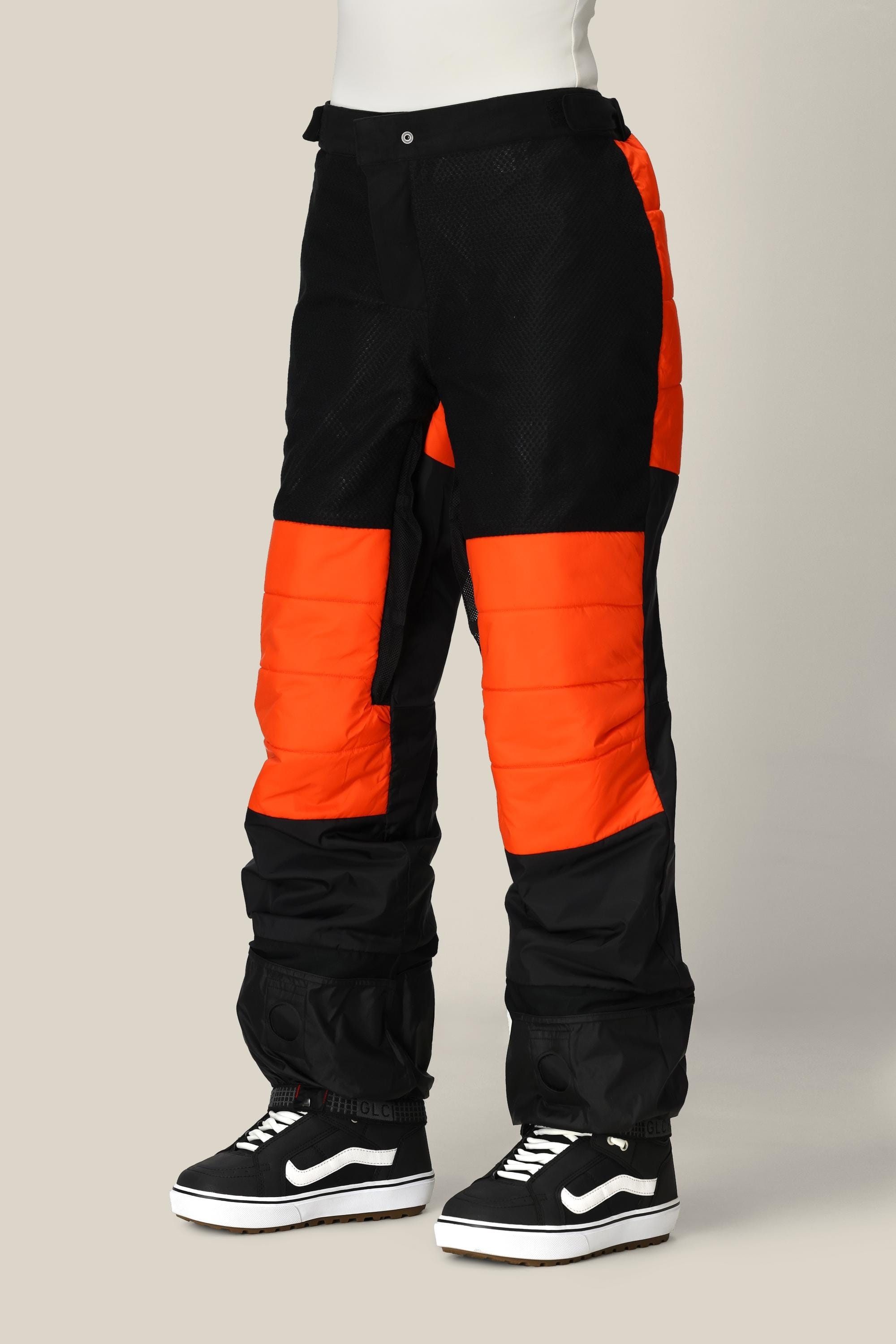 A person wearing the 686 Women’s Geode Thermagraph® Pant by 686 in black and orange, featuring waterproof polyester material and padded knees, paired with black and white Vans sneakers, stands against a plain beige background. Only the lower half of the body is visible.