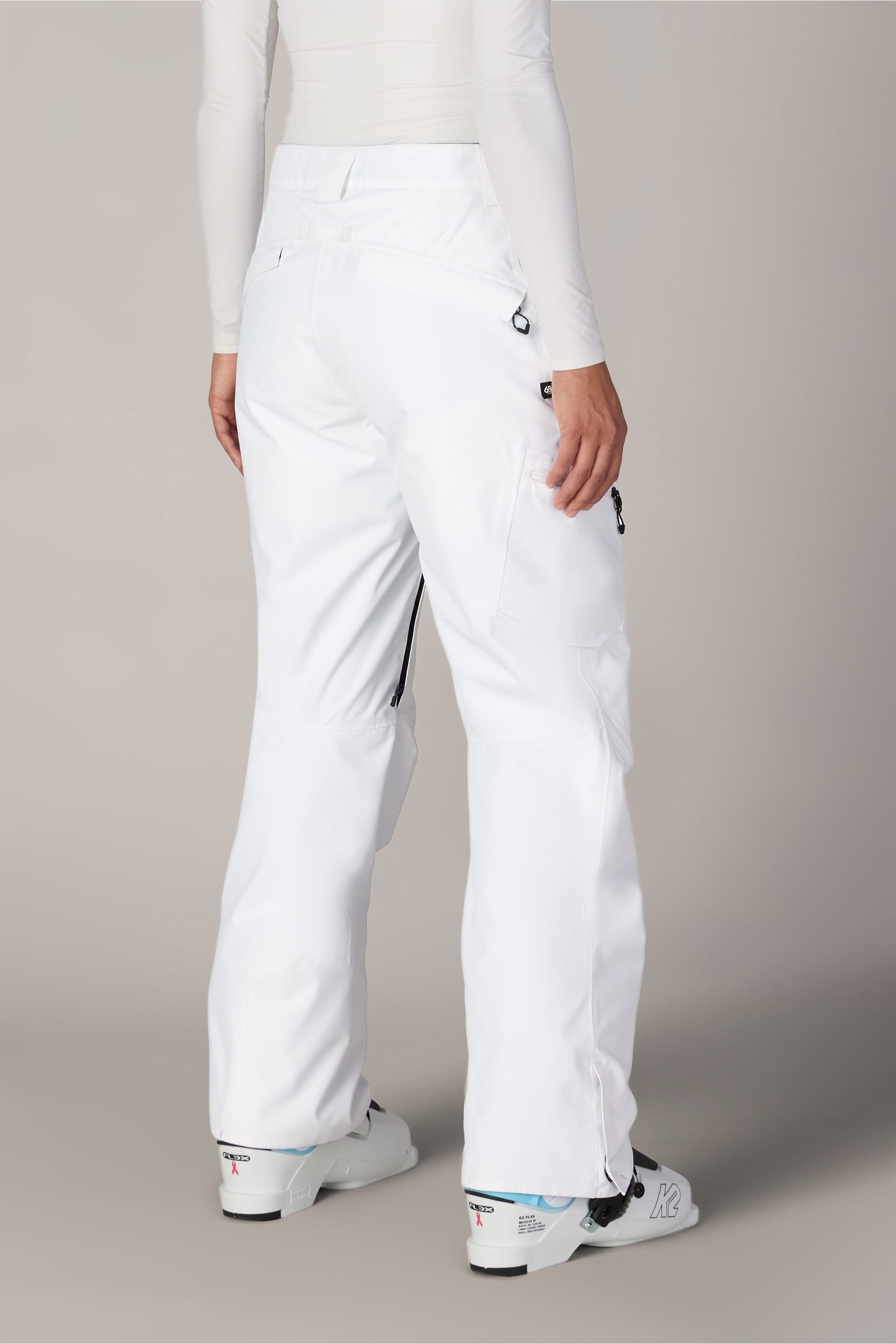 A person is shown from the back, with only their lower body visible, wearing the 686 Women’s Geode Thermagraph® Pant by 686. The white, waterproof polyester pants feature zippered pockets and black accents and are paired with white ski boots, set against a neutral background.