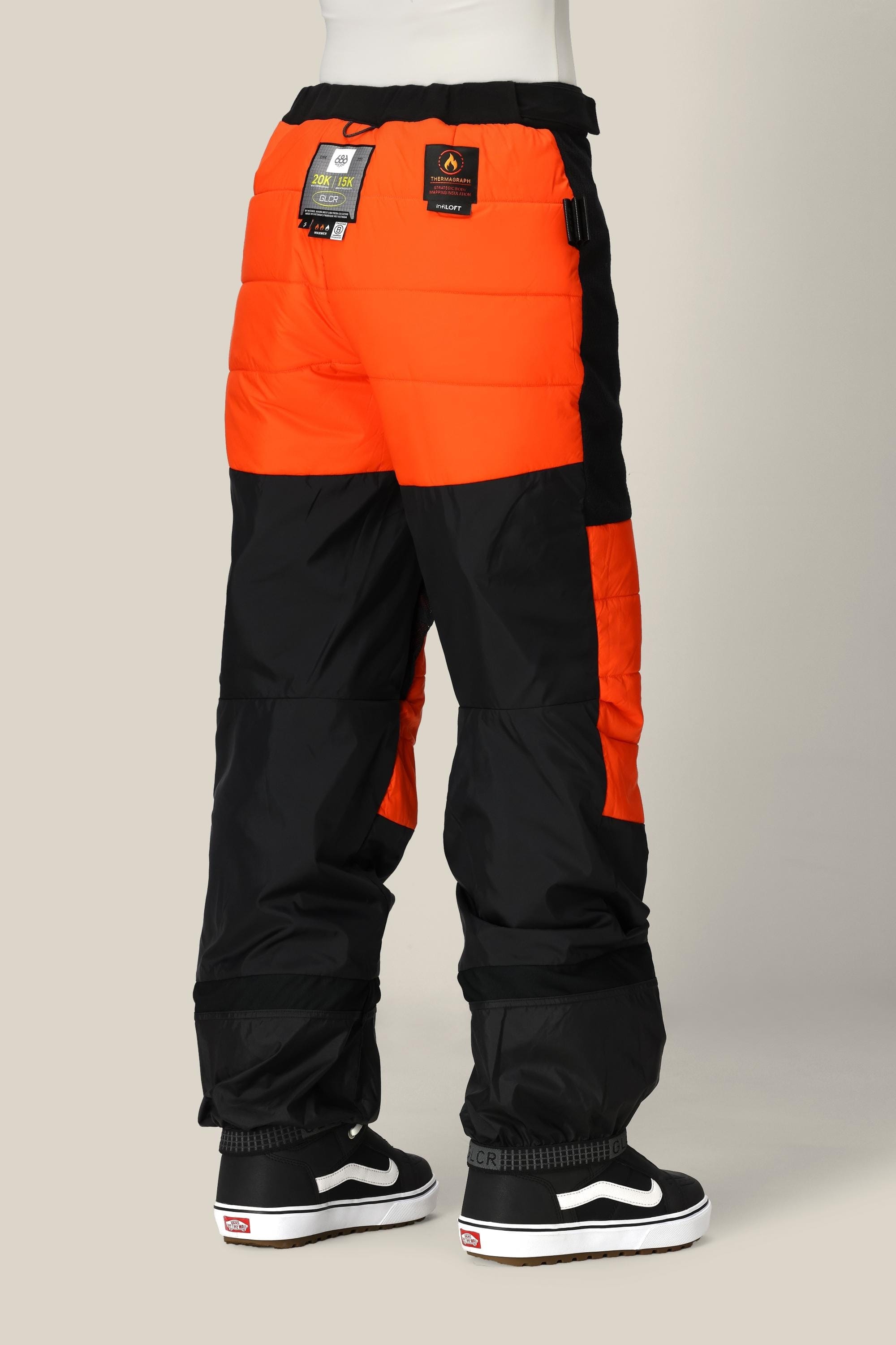 A person wearing the 686 Women’s Geode Thermagraph® Pant—black and orange color-blocked waterproof polyester pants by 686, featuring labeled waistband details—paired with black and white Vans sneakers, stands against a plain light background.