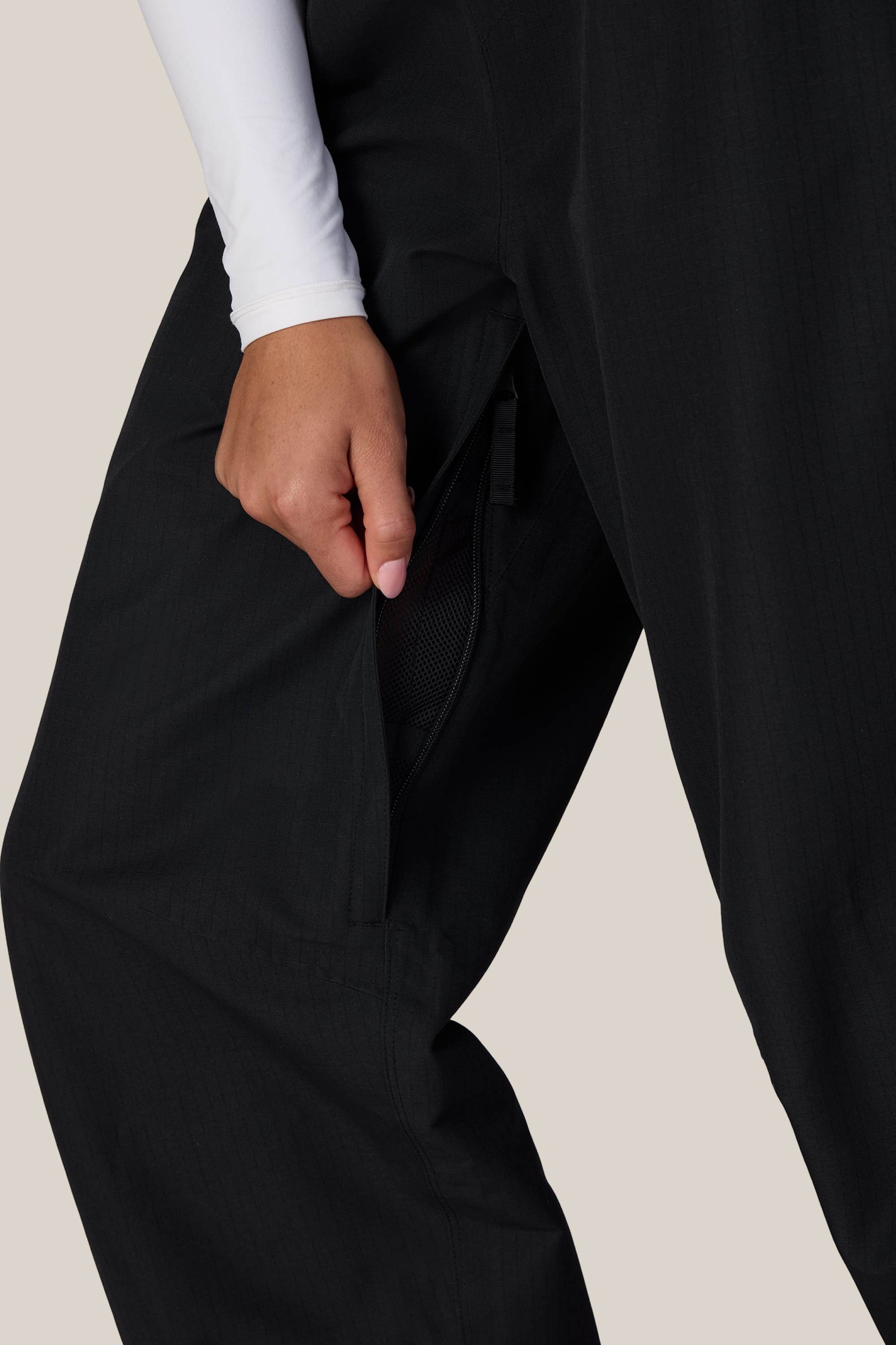 A person wearing 686 Women’s Hellkat™ Shell Pants in black and a white long-sleeve shirt opens a zippered side pocket, revealing the mesh lining inside.