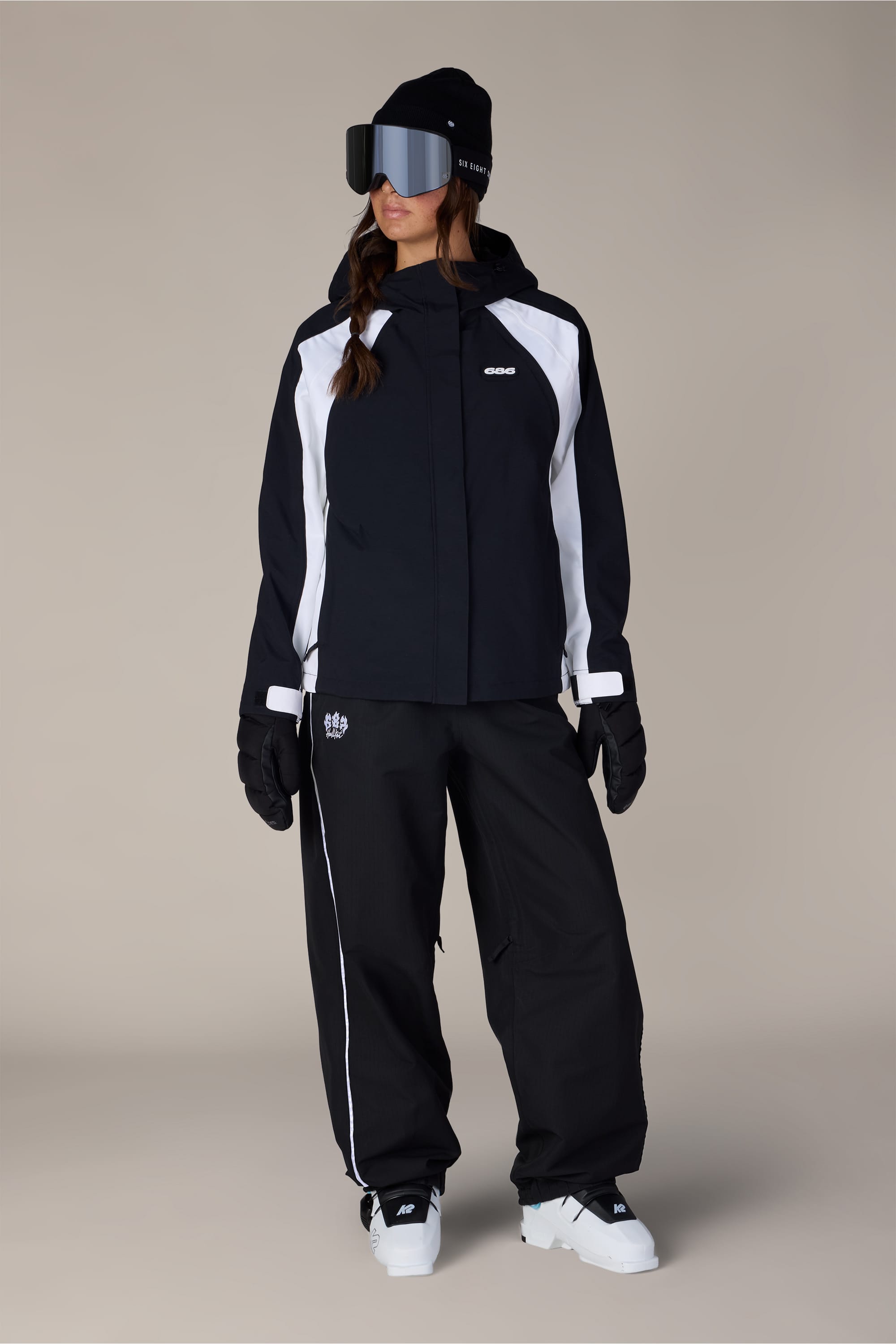 A person stands facing left, wearing a black and white ski outfit featuring the 686 Women’s Hellkat™ Shell Pant by 686, large reflective ski goggles, black gloves, and white boots. The background is plain and neutral.