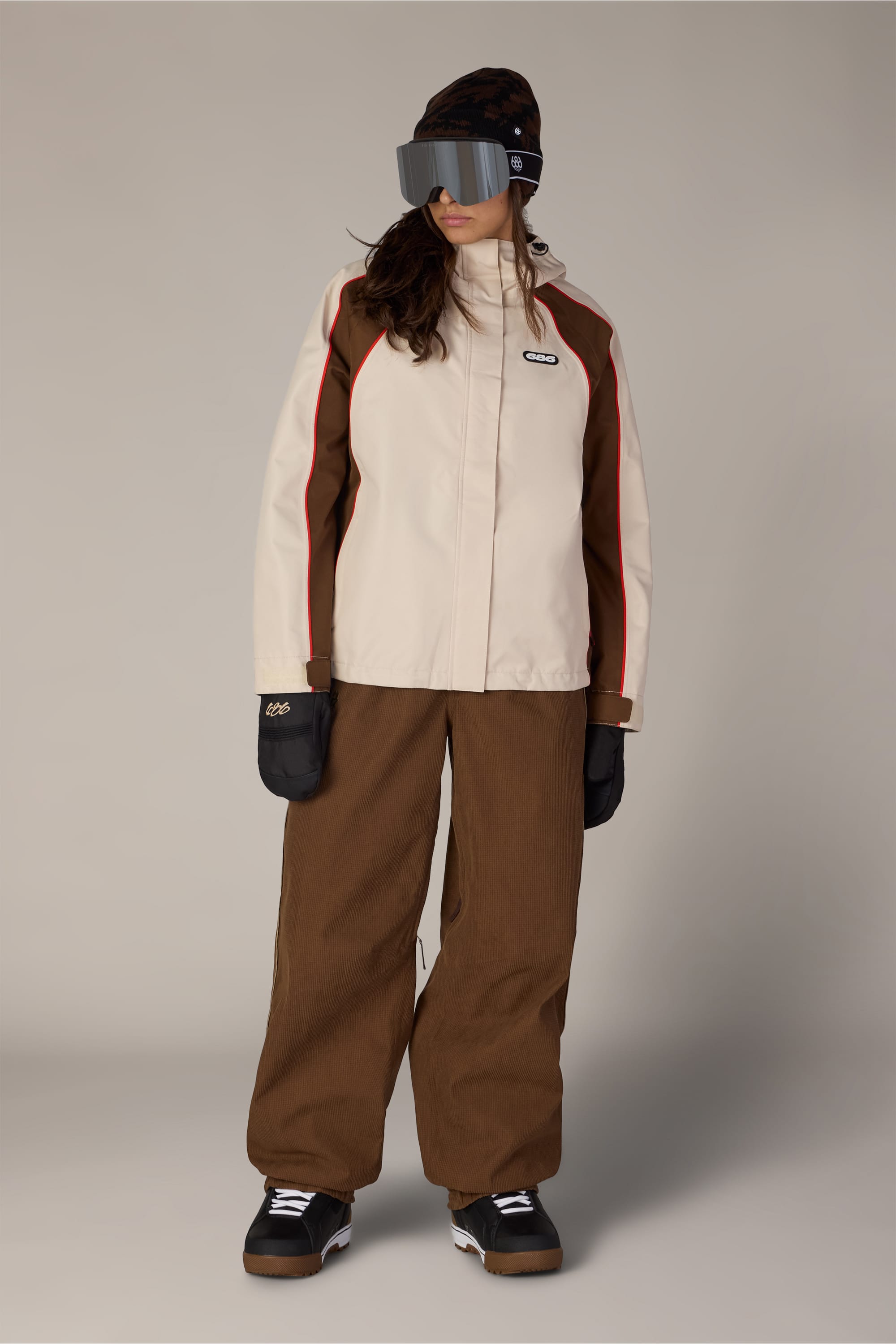 A person wearing a beige and brown ski jacket, 686 Women’s Hellkat™ Shell Pant by 686, black gloves, black boots, a black beanie, and large ski goggles stands against a plain background.
