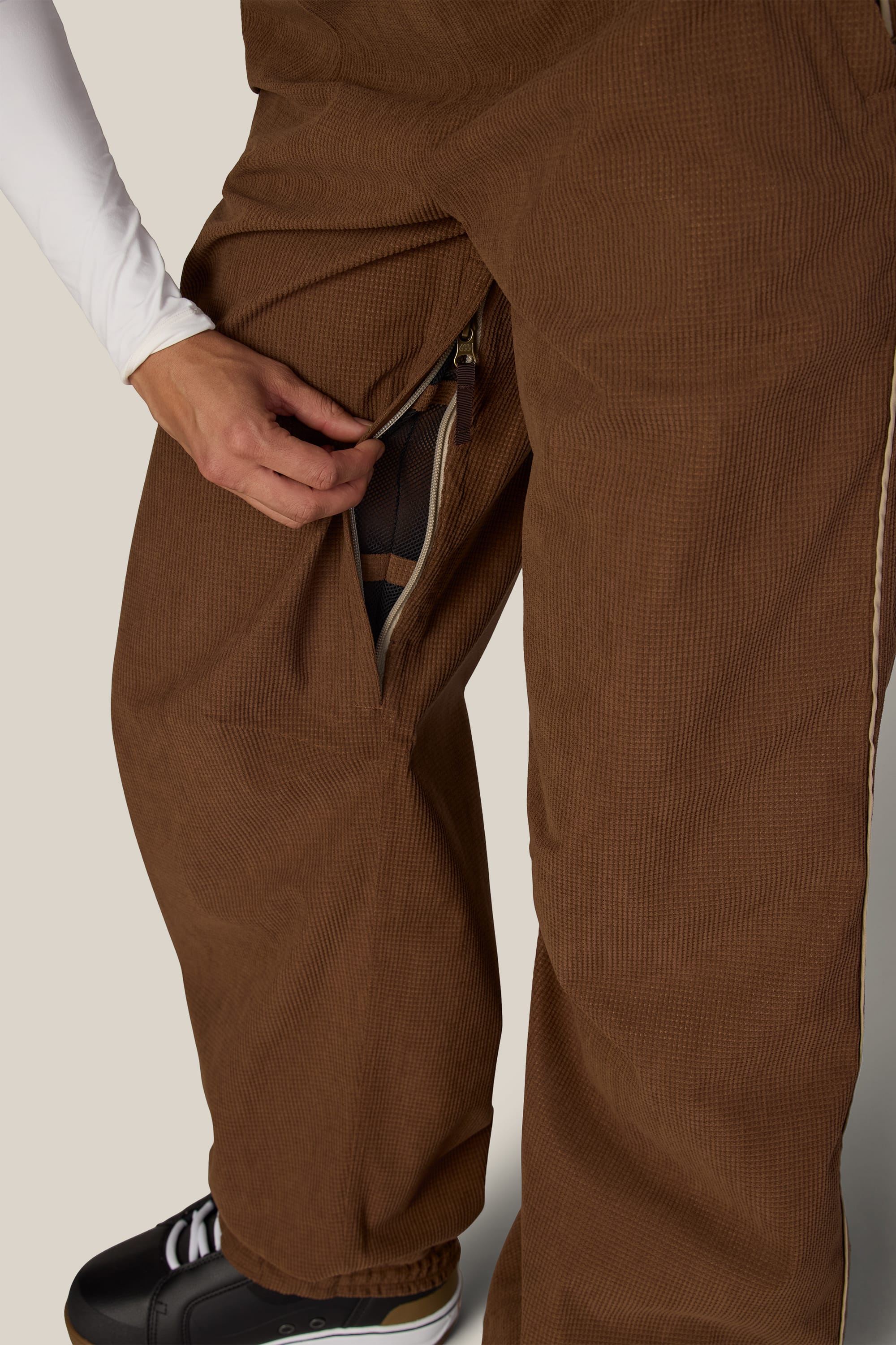 A person wearing the 686 Women’s Hellkat™ Shell Pant in brown and black sneakers unzips a side vent to reveal the mesh lining inside the pants. Only their hand and lower body are visible.