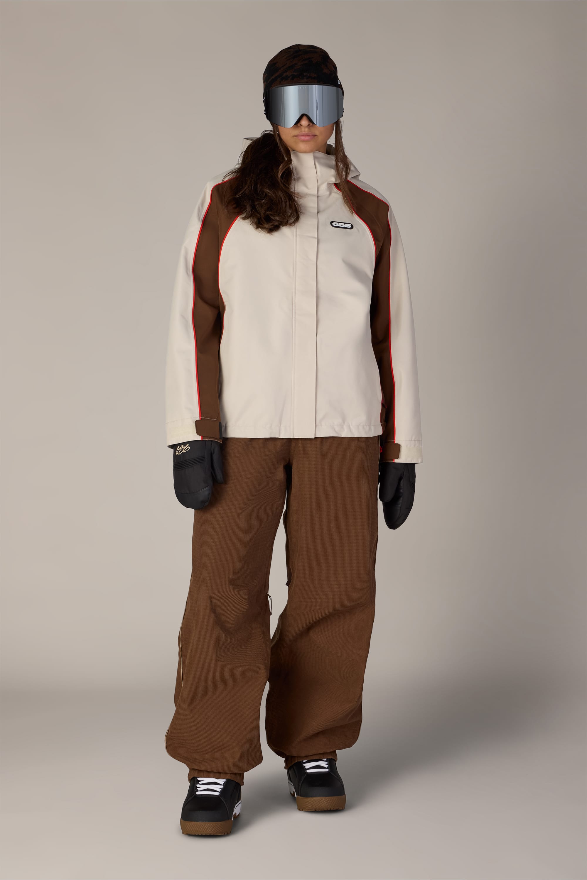 A person stands facing forward against a plain background, wearing a beige and brown ski jacket, black gloves, black shoes, large ski goggles, and 686 Women’s Hellkat™ Shell Pant in brown by 686.