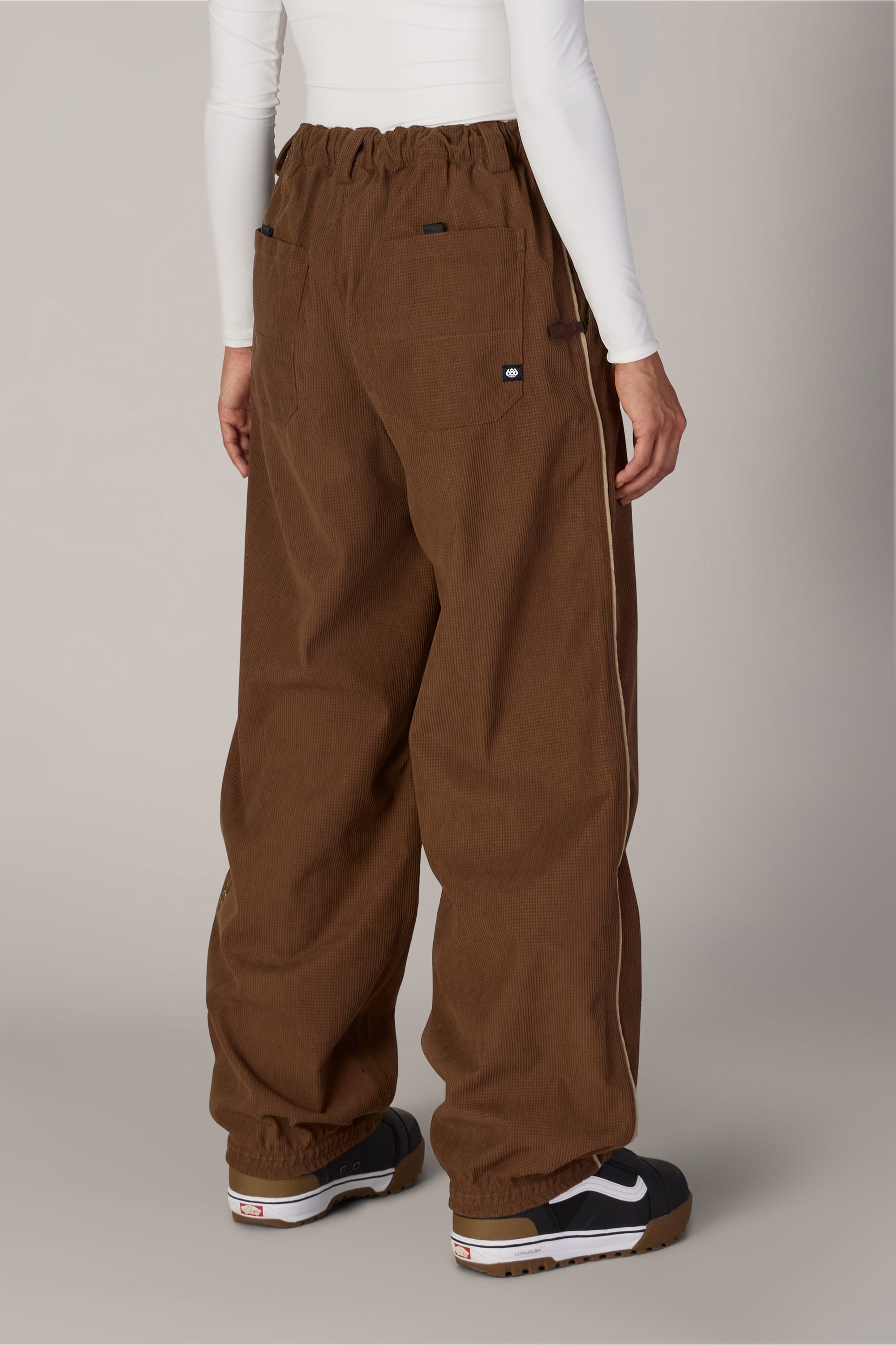 A person is shown from the back standing against a plain background, wearing 686 Women’s Hellkat™ Shell Pants by 686 styled like women’s woven pants, paired with a white long-sleeve shirt and black Nike sneakers.