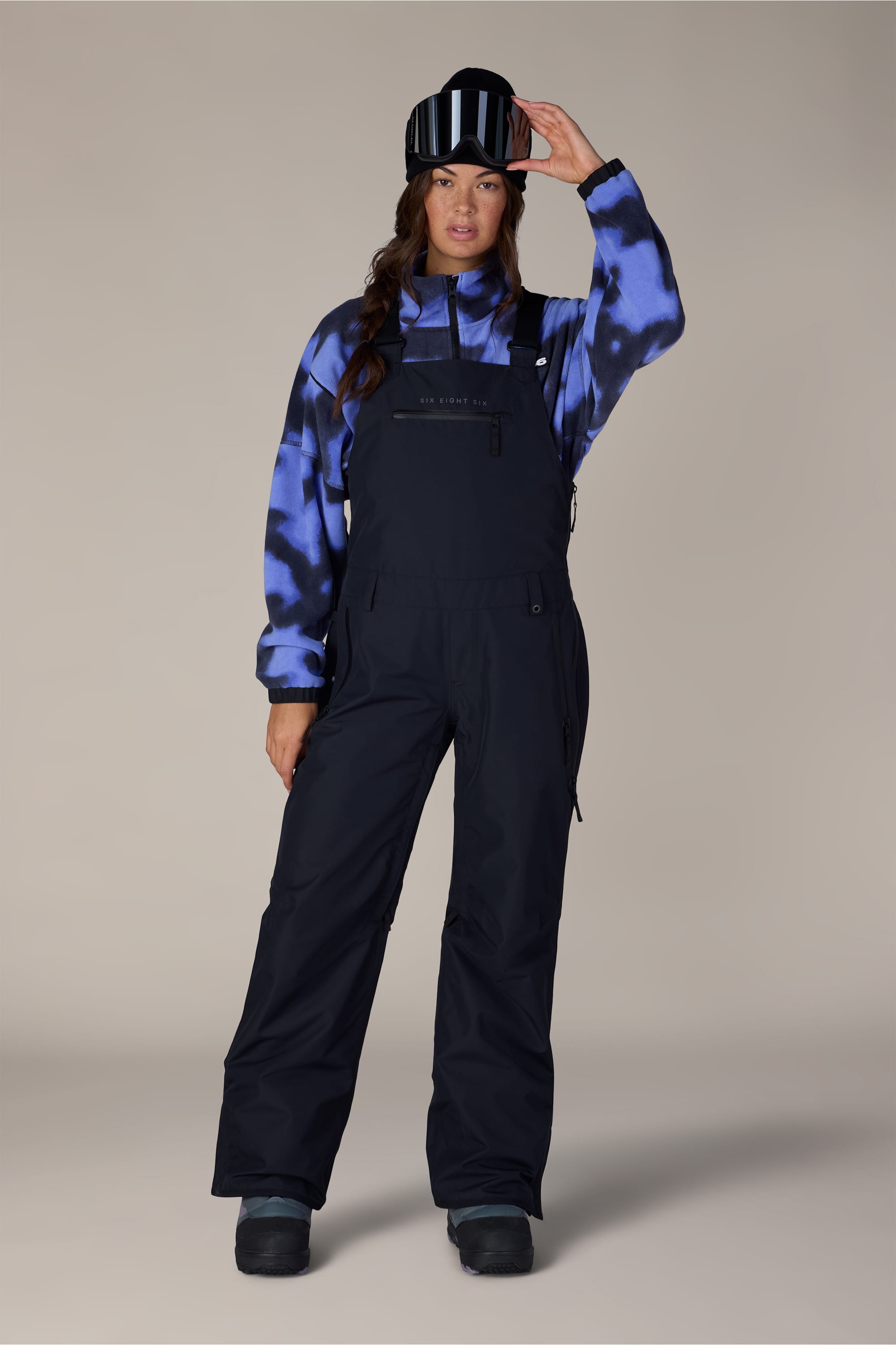 A person wearing a blue and black tie-dye hoodie, the 686 Women’s Regalia™ Bib snow overalls in black from 686, black snow boots, and holding a black ski helmet with white stripes against a plain background.