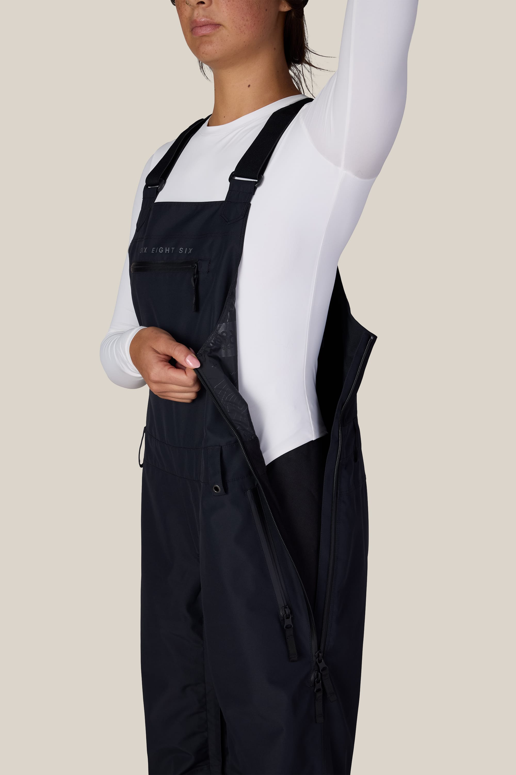 A person wearing the 686 Women’s Regalia™ Bib in black, styled with a white long-sleeve shirt, unzips the side zipper to reveal the inner lining. Their face is partially out of frame.