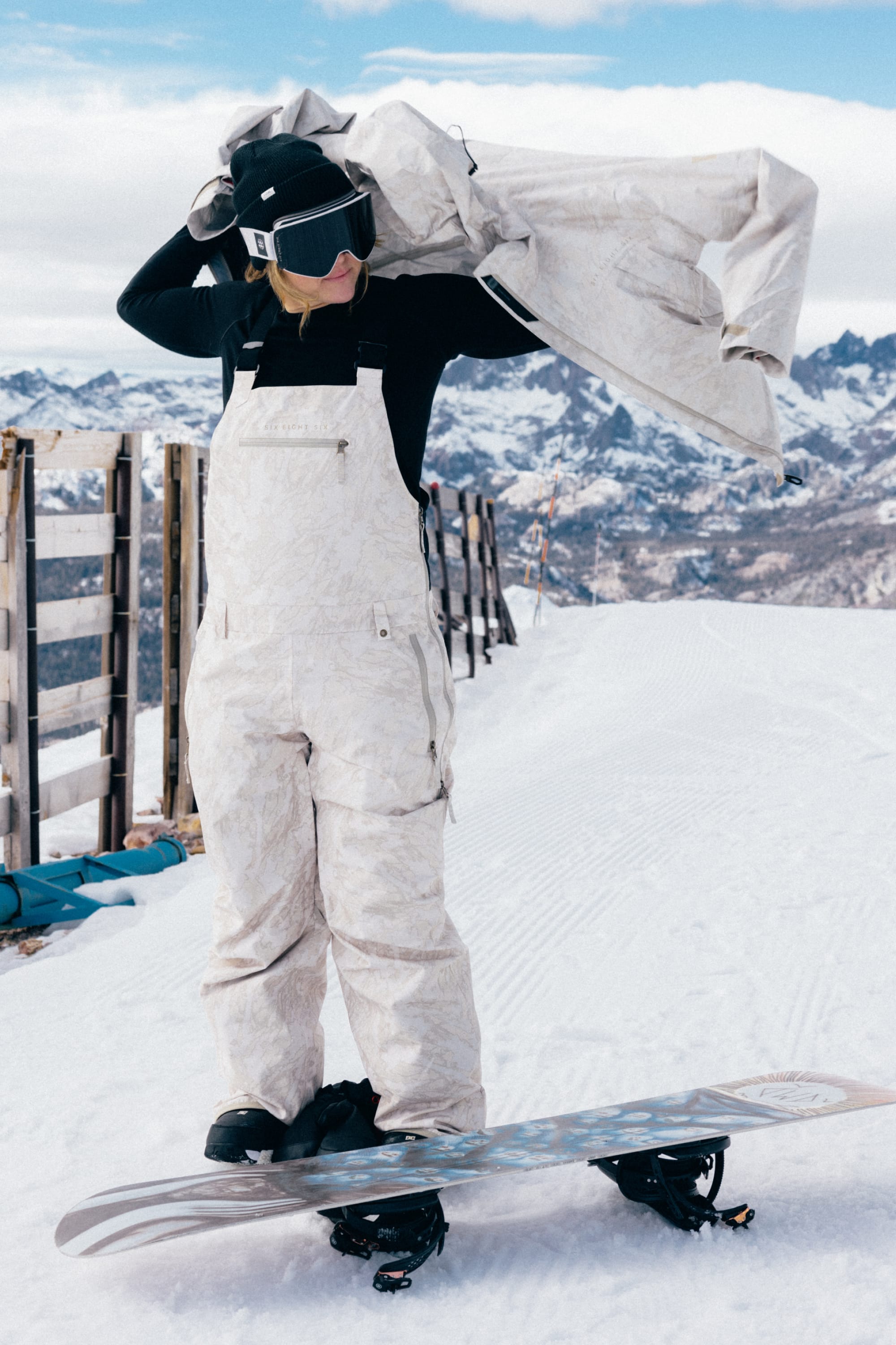 Wearing the 686 Women’s Regalia™ Bib, a person in white snow gear and goggles stands on a snowboard, adjusting their jacket with snowy mountains and a wooden fence in the background.