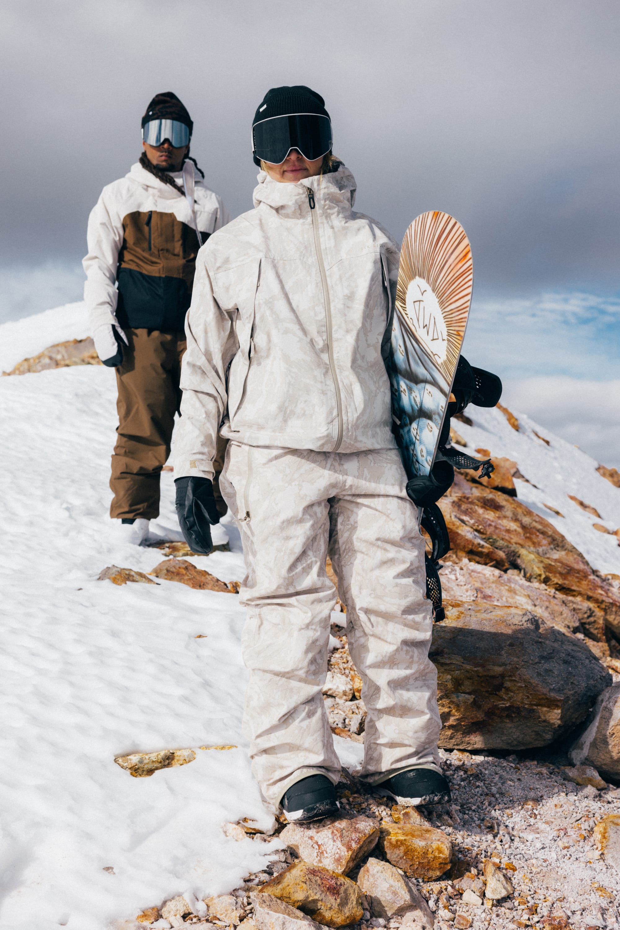 Clad in the 686 Women’s Regalia™ Bib for extra protection against the elements, two people in winter gear stand on a snowy, rocky slope. The person in front holds a snowboard, and both wear goggles, gloves, and hoods. With cloudy skies overhead, they look ready to take on the wintry cold—thanks to their waterproof bibs from 686.