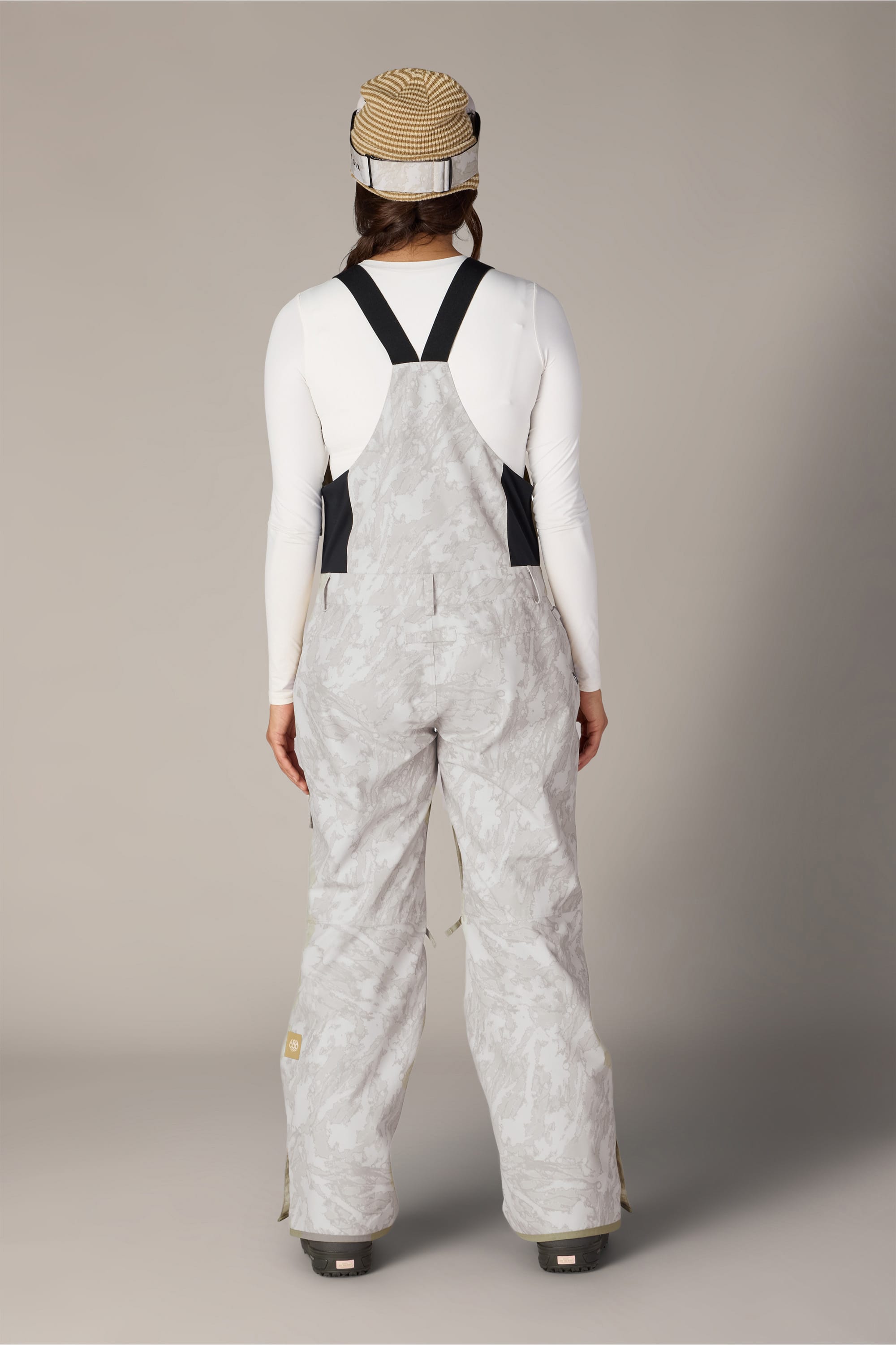 A person stands facing away, wearing a striped beanie, a white long-sleeve shirt, and the 686 Women’s Regalia™ Bib—light gray camouflage waterproof overalls with black straps and black side panels by 686—against a plain backdrop.