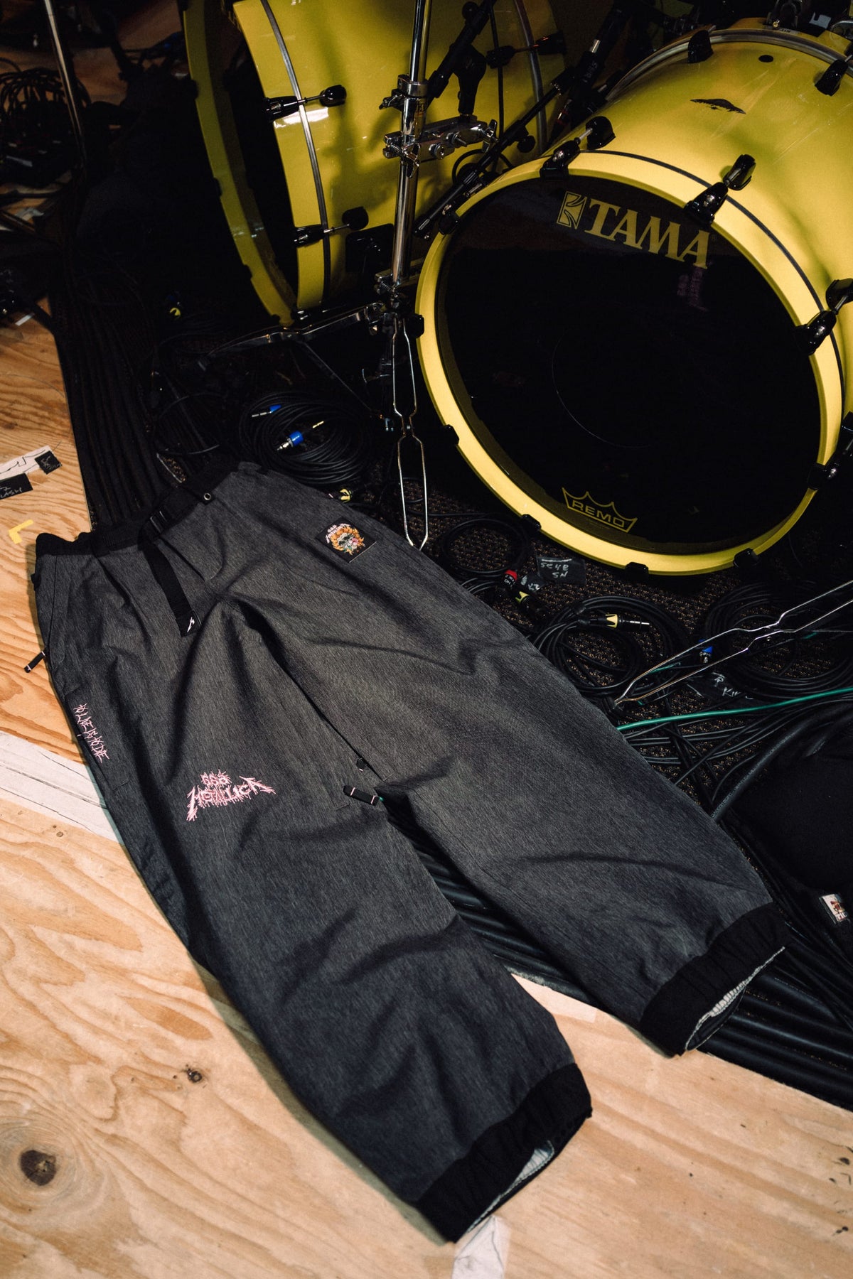 686 Women’s Metallica Burning Skull Shell Pant in dark grey, featuring a pink Metallica logo and skull patch, laid on a floor near a yellow bass drum.