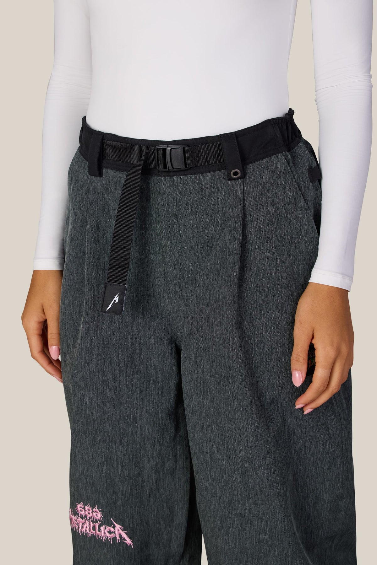Dark grey waterproof woven 686 Women’s Metallica Burning Skull Shell Pant featuring a black webbing belt and a pink Metallica logo on the lower leg.