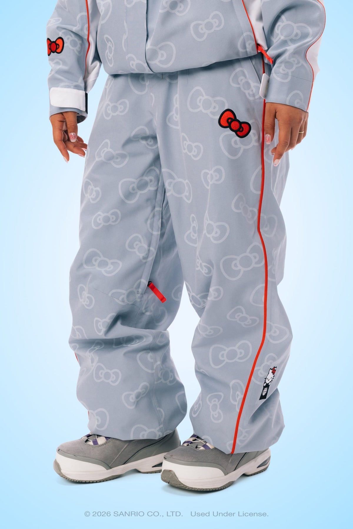 A person is wearing the 686 Women’s Hello Kitty® Lil Bow Shell Pant by 686, a light blue track pant featuring white outlines of Hello Kitty’s face and red accents, paired with gray sneakers, and standing against a light blue background. Only the lower half of the body is visible.
