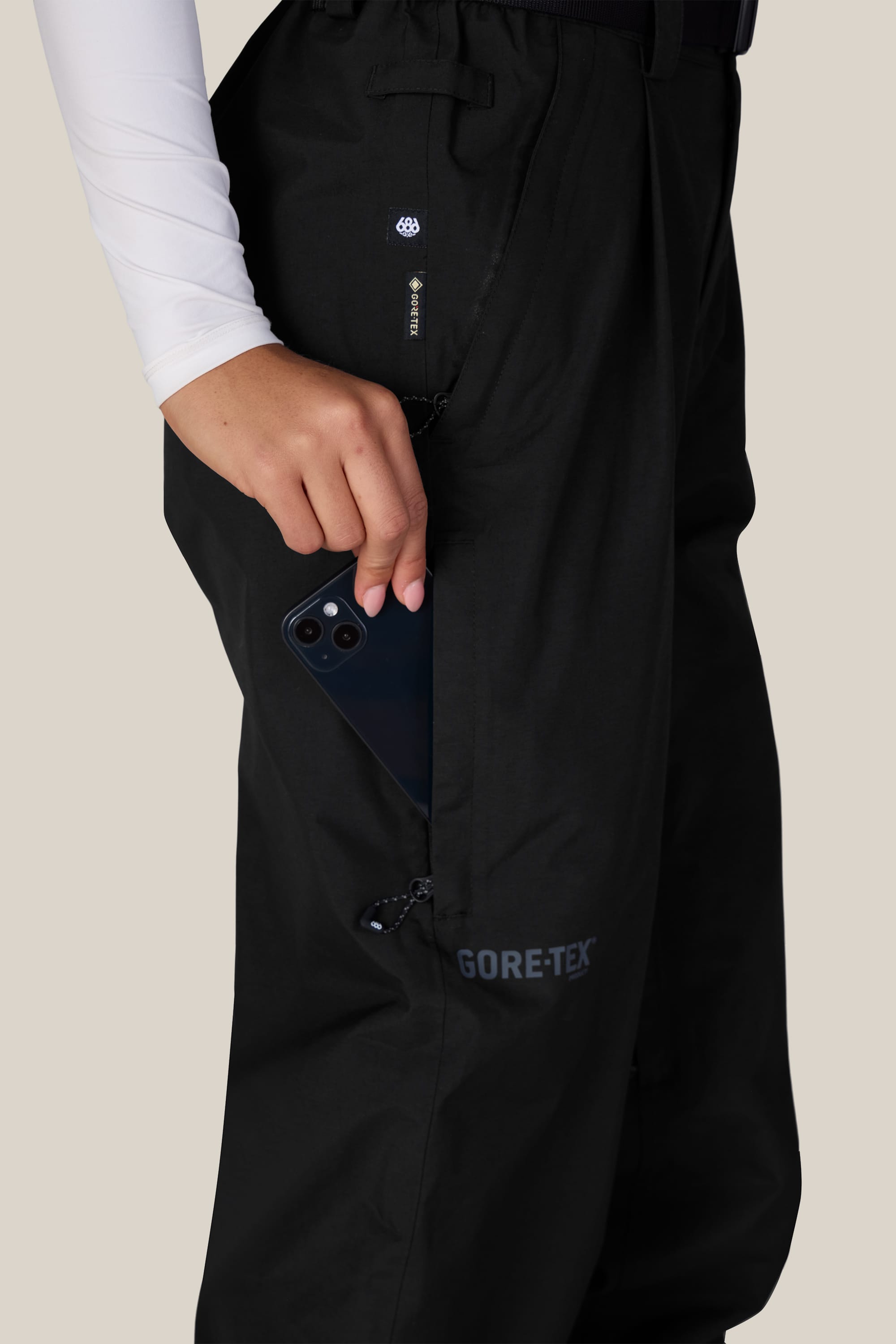 A person wearing the 686 Women’s GORE-TEX® Outline™ Pant in black and a white long sleeve shirt puts a black smartphone into a zippered side pocket.