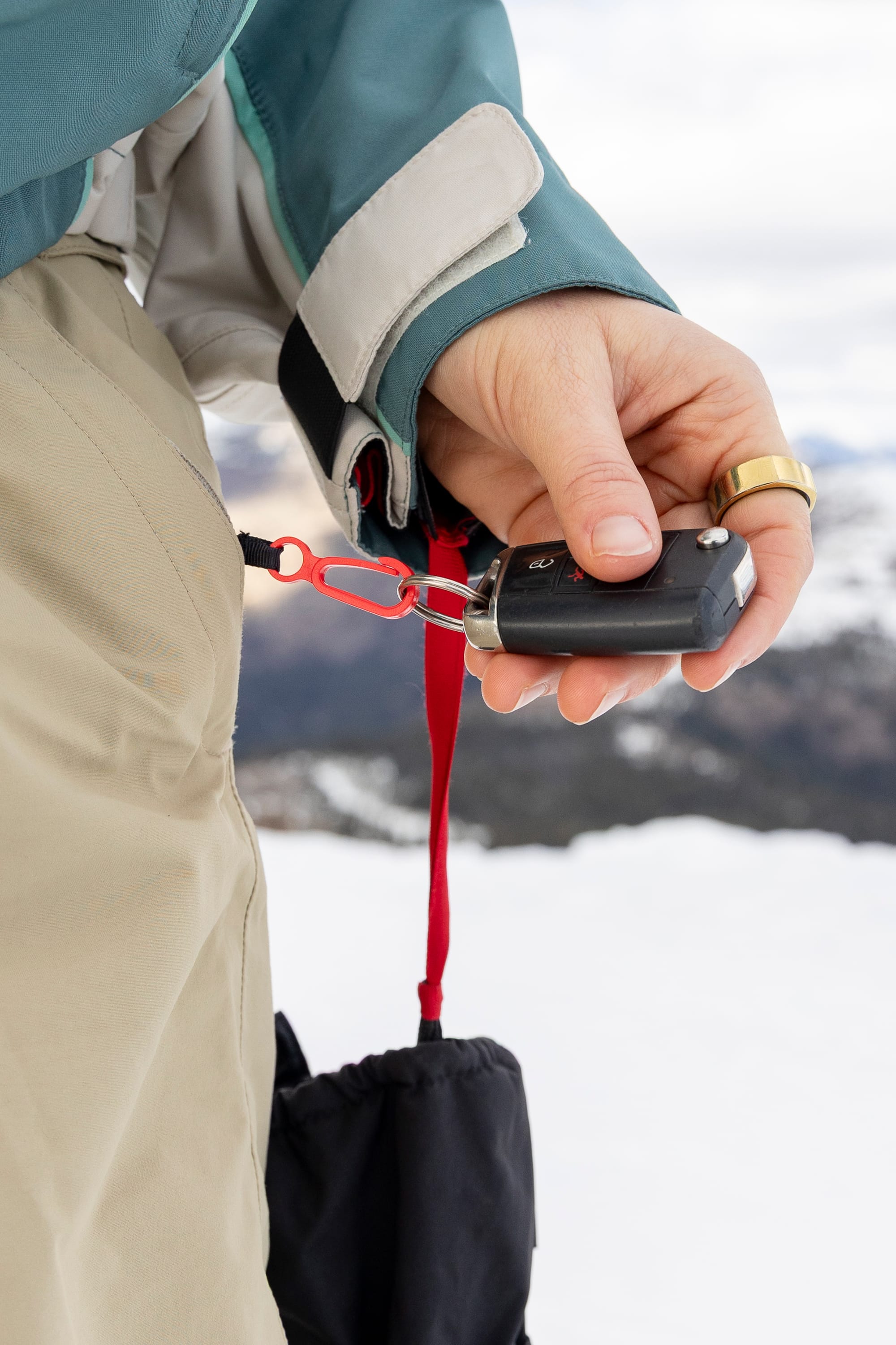 Wearing the 686 Women’s GORE-TEX® Outline™ Pant in beige, a person stands outdoors in a snowy landscape, securing a black device to their pants with a red carabiner and strap.