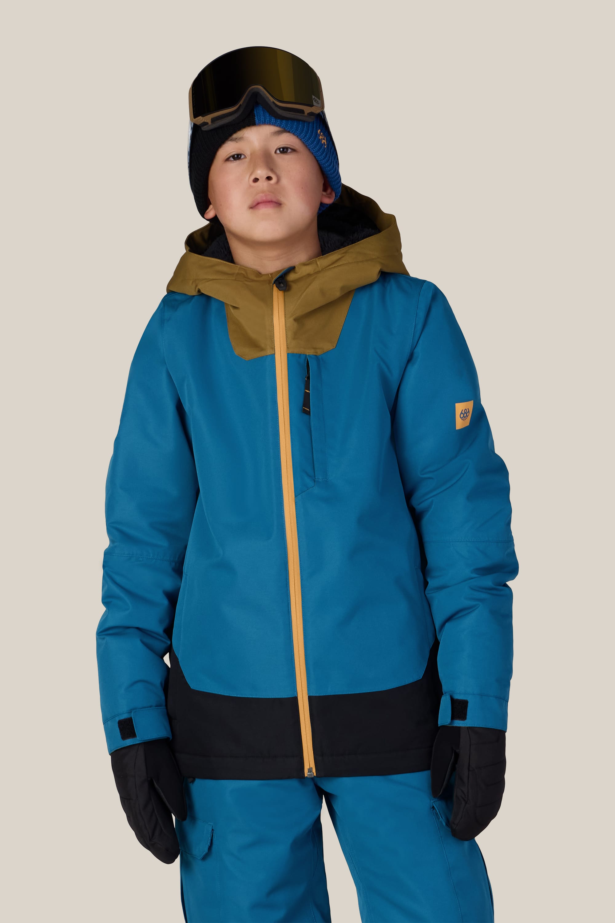 A child wearing the 686 Boys’ Gateway™ Insulated Jacket by 686 in blue and brown, along with blue snow pants, black gloves, a dark blue beanie, and ski goggles on their forehead, stands against a plain light background.