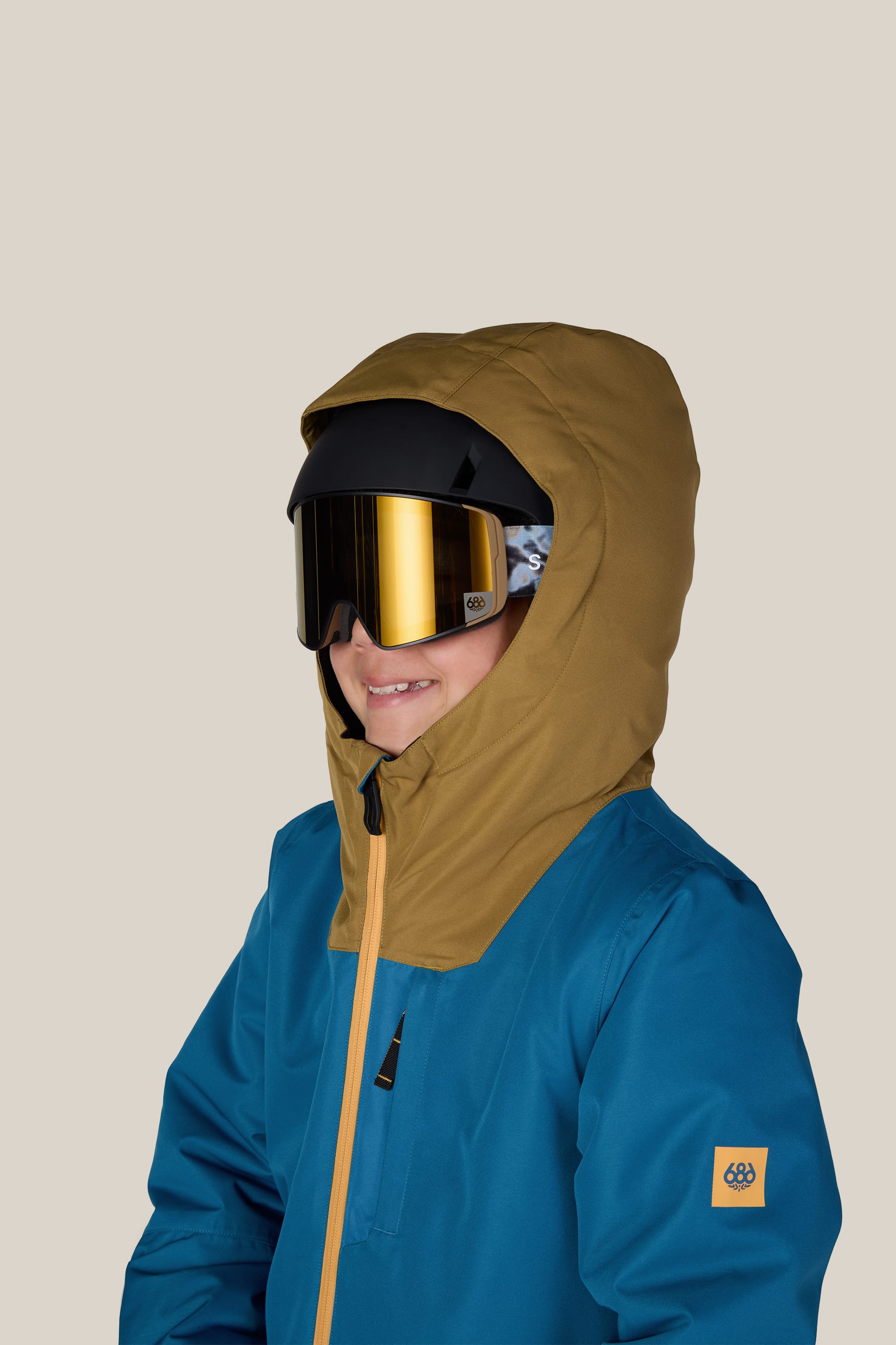 A person wearing the 686 Boys’ Gateway™ Insulated Jacket in blue, featuring a brown hood and reflective ski goggles, smiles against a plain light background.