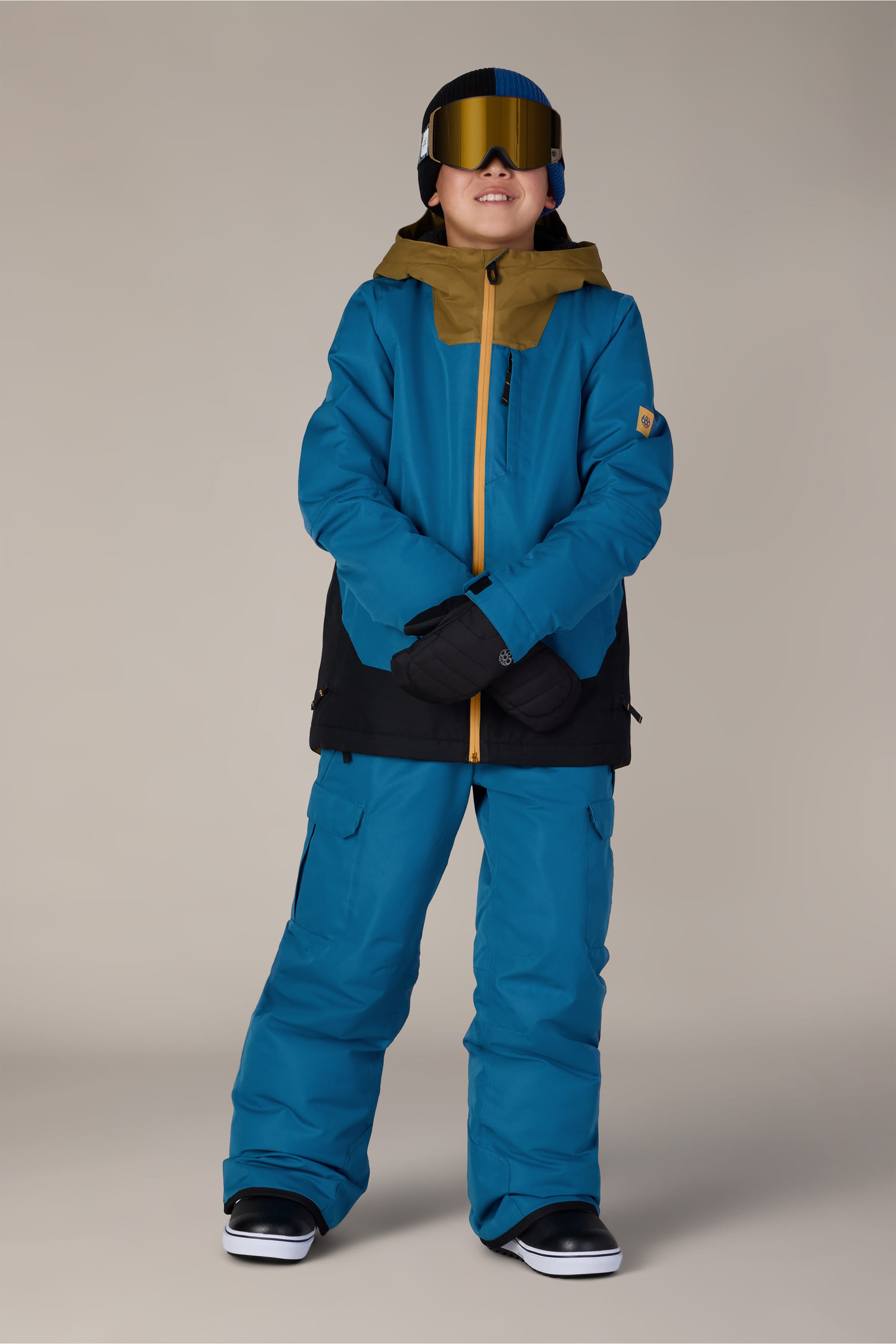 A child wearing the 686 Boys’ Gateway™ Insulated Jacket in blue and black, along with blue snow pants, black gloves, black snow boots, a gold and black hood, and ski goggles, stands smiling against a plain background.