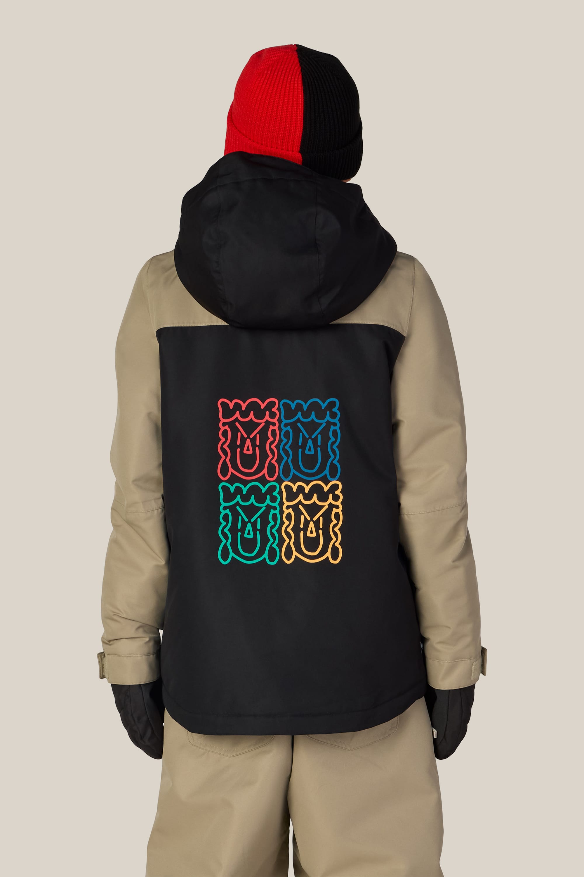 A person faces away wearing the 686 Boys’ Gateway™ Insulated Jacket by 686, featuring a beige and black woven design with colorful abstract face artwork on the back. They pair the jacket with a red and black beanie, black gloves, and tan pants, standing against a plain light background.