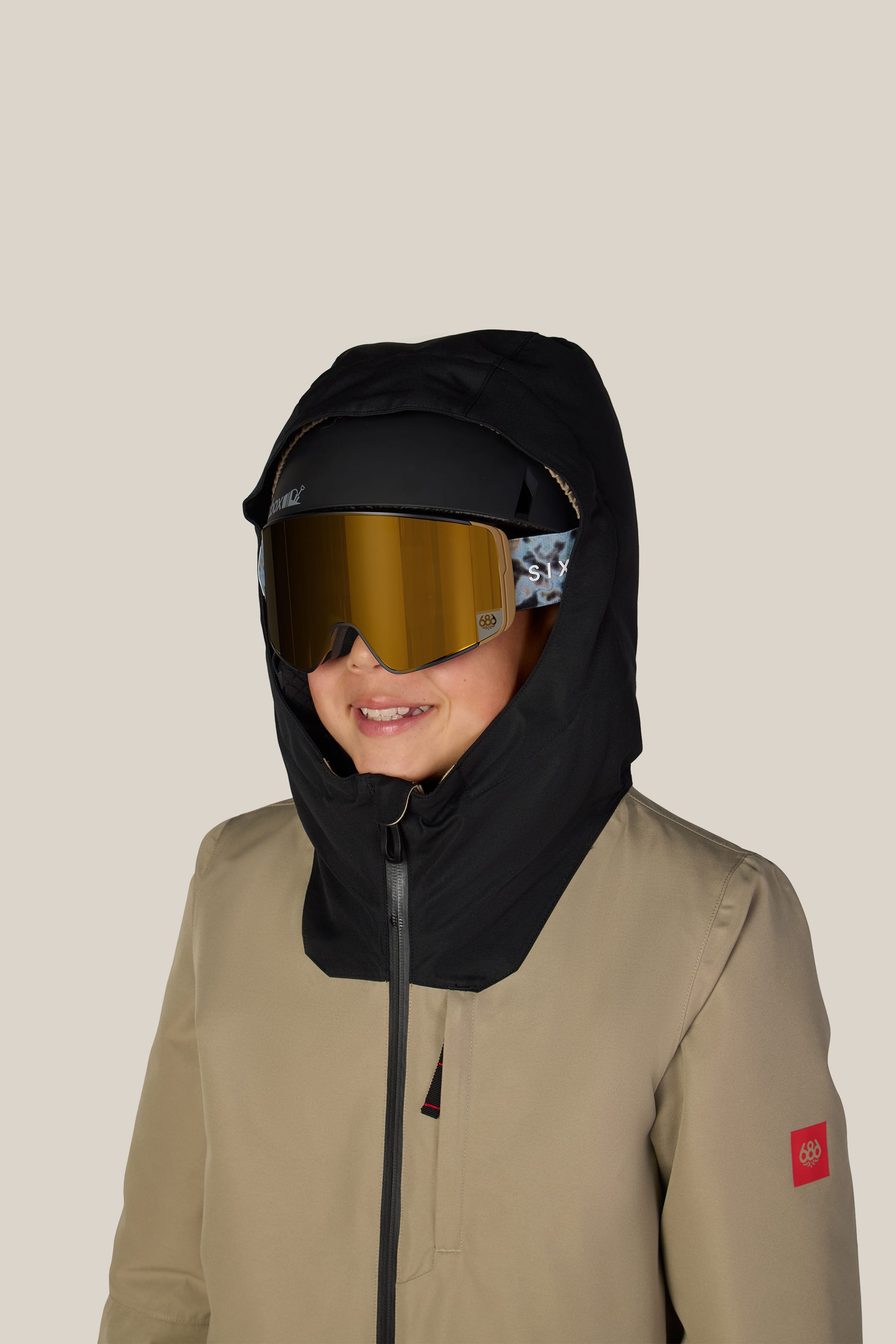 A child wearing the 686 Boys’ Gateway™ Insulated Jacket in tan, paired with a black hood, gold reflective ski goggles, and a black helmet, smiles against a plain light background.