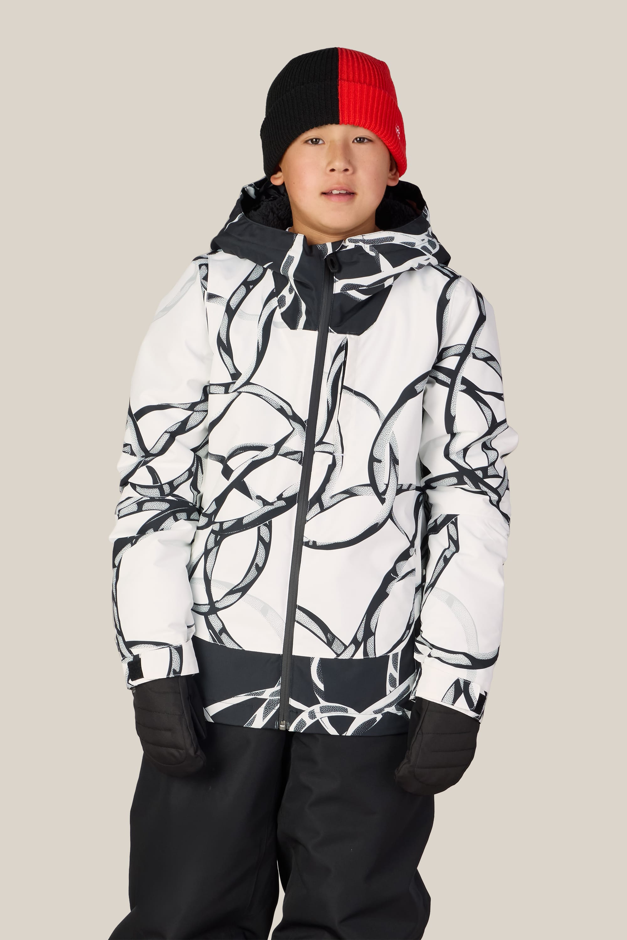 A child wearing the 686 Boys’ Gateway™ Insulated Jacket by 686, featuring a white and black pattern, along with black gloves, black pants, and a red and black beanie stands against a plain light background.