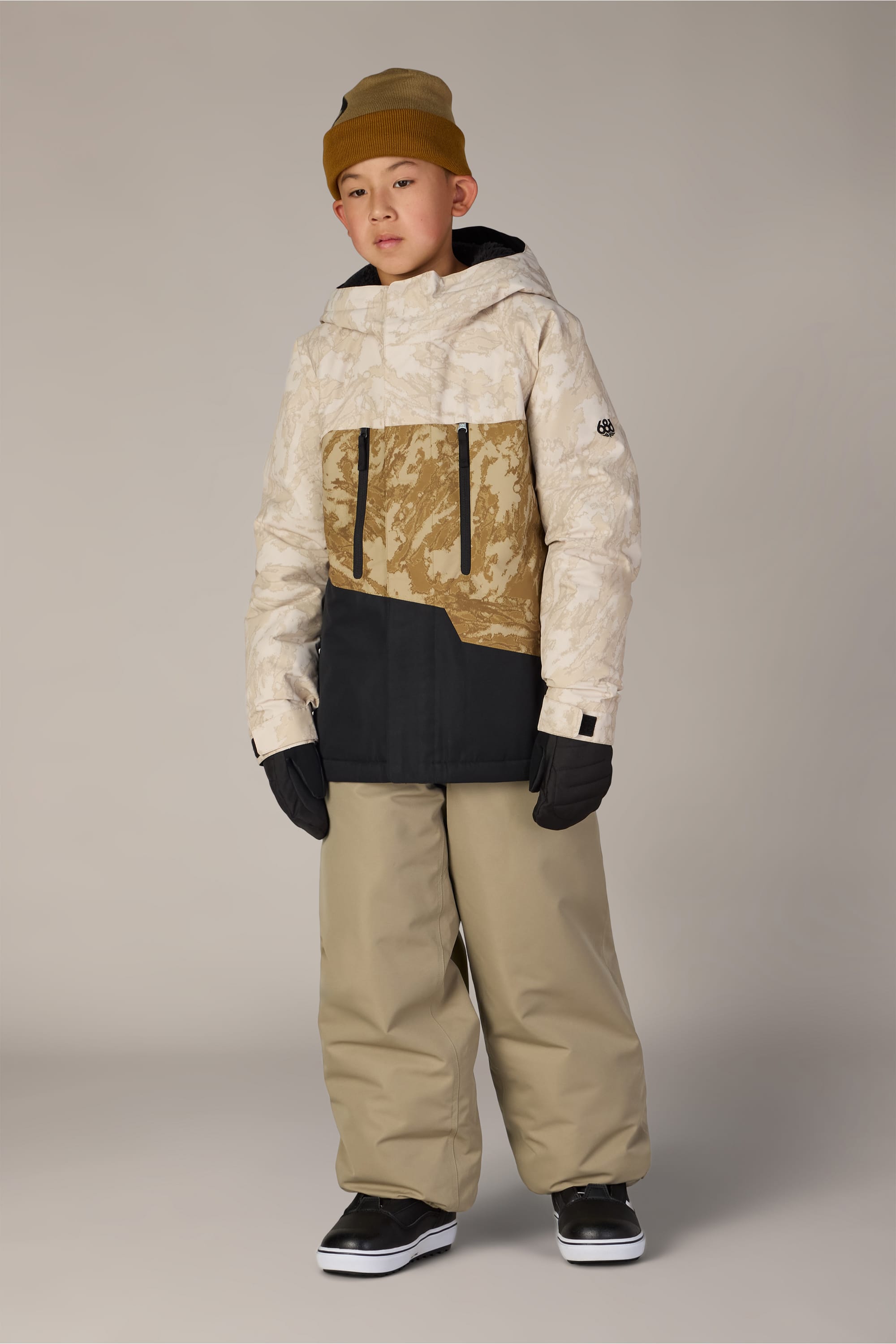 A young person wearing a tan beanie, a patterned tan and white jacket, black gloves, 686 Boys’ 686 Dojo® Insulated Pant by 686 in a baggy khaki style, and black sneakers stands against a plain background.